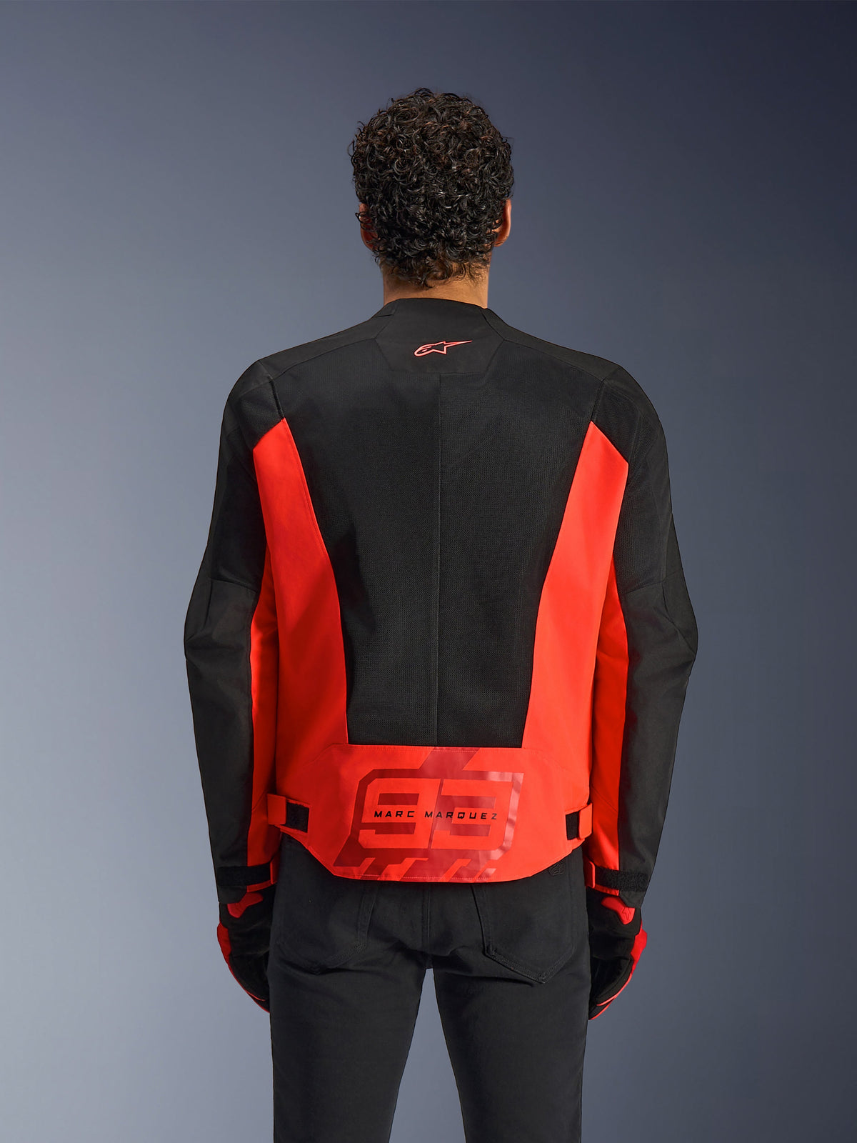 Vista posteriore di un uomo che indossa la giacca Alpinestars MM93 C-1 Air in Rosso Fluo e Nero, caratterizzata da un grande logo '93' di Marc Marquez sulla parte bassa della schiena, pannelli in mesh leggero per la ventilazione, abbinata a pantaloni e guanti neri.