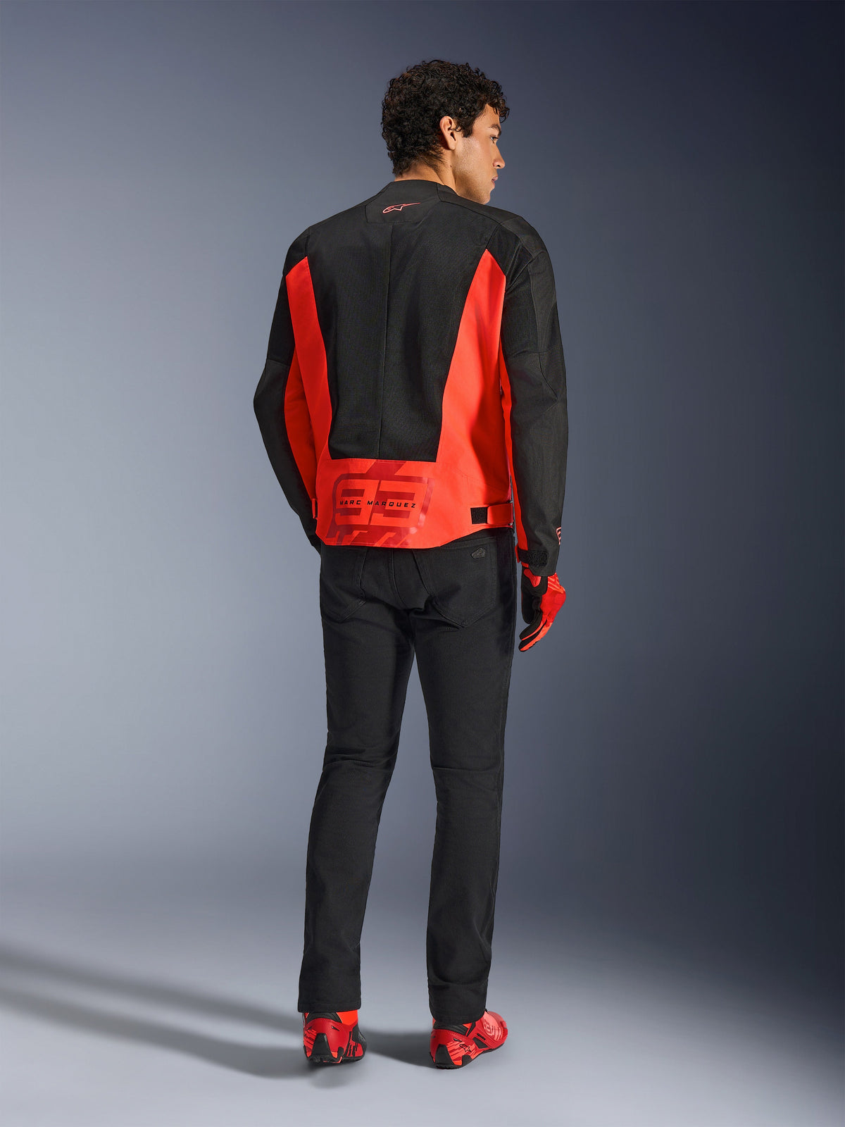 Vista posteriore di un modello che indossa la giacca Alpinestars MM93 C-1 Air in rosso fluo e nero, con il logo Marc Marquez 93 sulla parte inferiore della schiena, abbinata a pantaloni neri e scarpe da moto rosse, stile urban motorcycle commuting