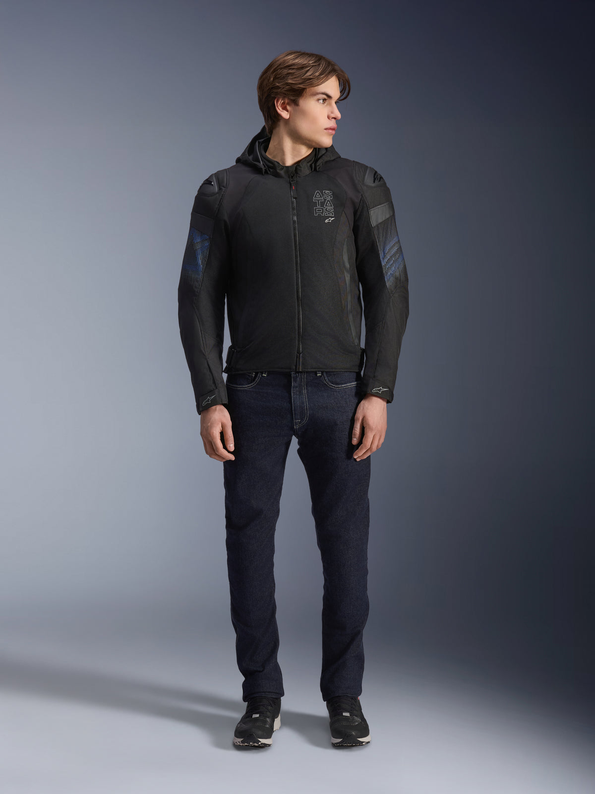 Modello che indossa la giacca Alpinestars Zaca Air Venom WP in nero e blu elettrico, giacca da moto in tessuto con pannelli in rete 3D e cappuccio rimovibile, abbinata a jeans in denim scuro, abbigliamento tecnico da guida di ispirazione sportiva moderna