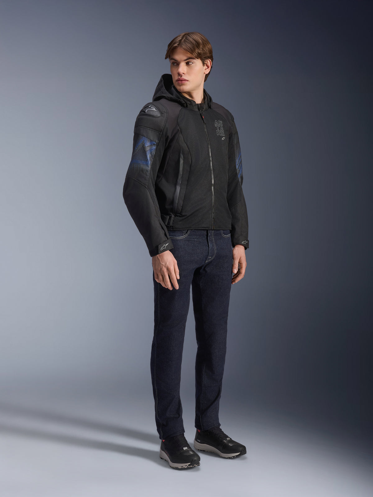 Giacca Alpinestars Zaca Air Venom WP, giacca da moto in tessuto nero e blu elettrico, indossata da un modello con jeans scuri e sneakers nere, dotata di cappuccio rimovibile, pannelli di ventilazione in mesh e slider sulle spalle DFS Lite, abbigliamento tecnico sportivo per tutte le condizioni atmosferiche.