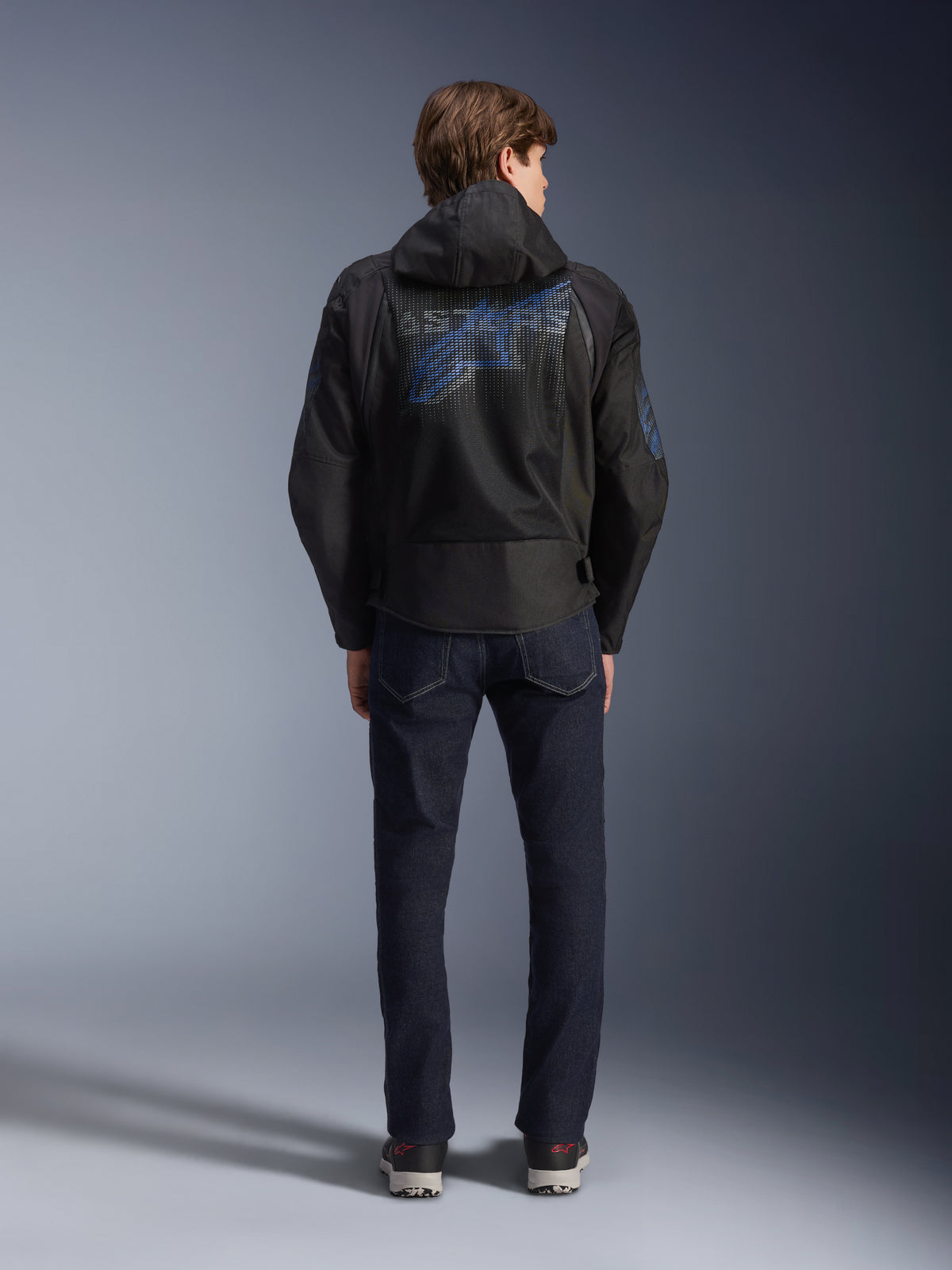 Giacca Alpinestars Zaca Air Venom WP, nero e blu elettrico, vista posteriore con un grande logo Alpinestars blu sul pannello posteriore in rete, cappuccio rimovibile, indossata con jeans in denim scuro e sneakers, abbigliamento da moto sportivo per tutte le stagioni.