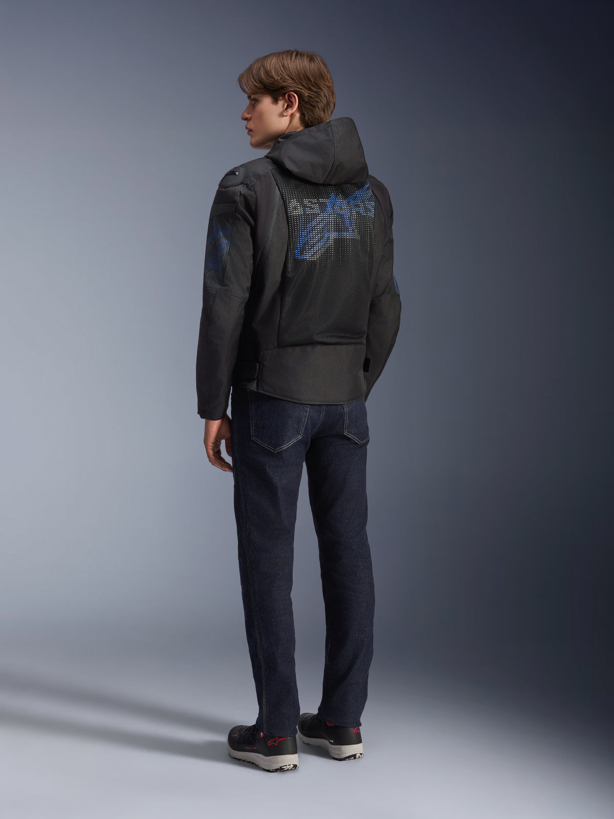 Giacca Alpinestars Zaca Air Venom WP, nero e blu elettrico, vista posteriore su un modello, giacca da moto in tessuto mesh con logo Astar blu sulla schiena, dotata di cappuccio rimovibile, indossata con jeans scuri e scarpe da moto nere.