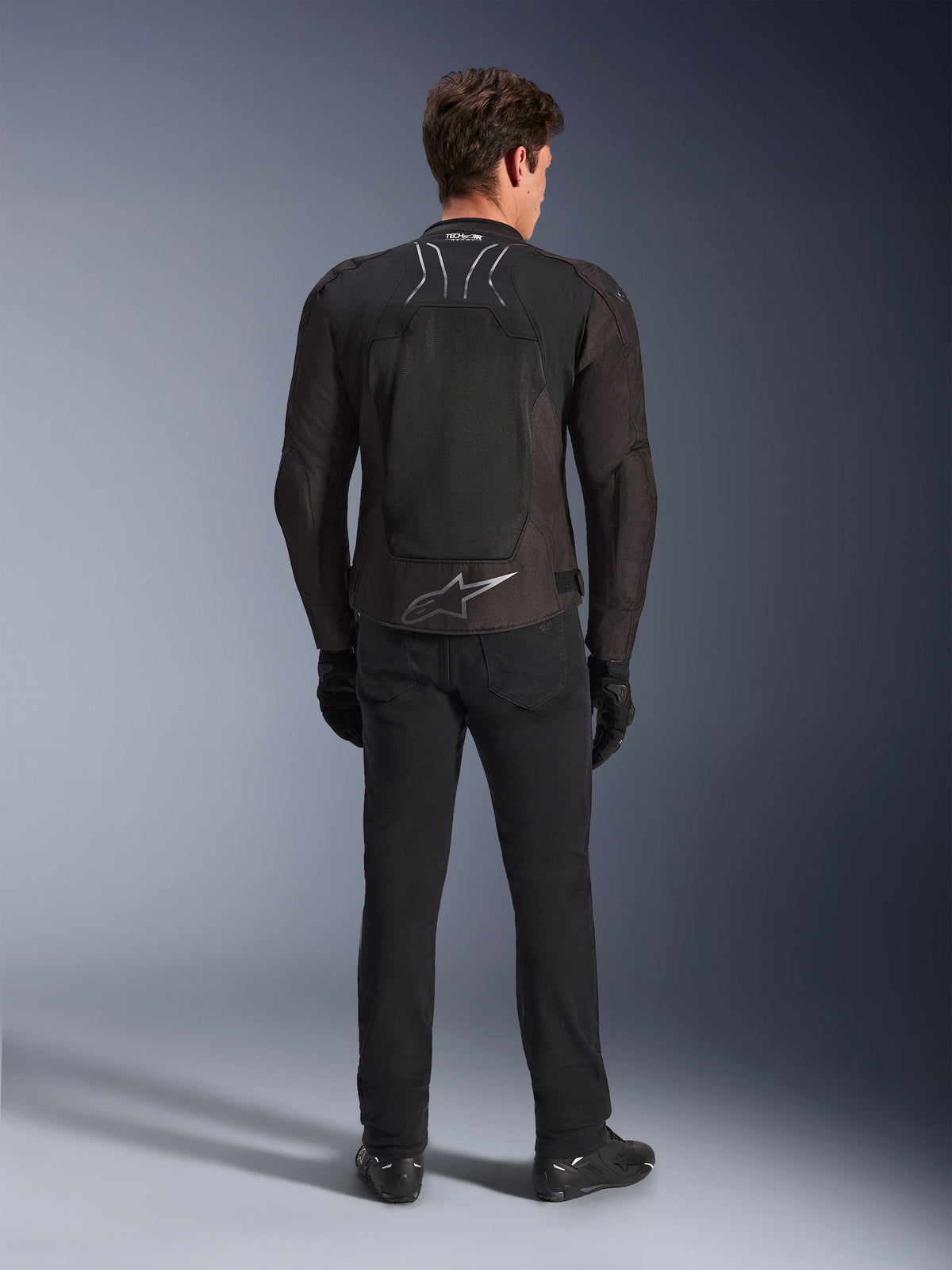 Giacca Alpinestars SMX Air, nero antracite, vista posteriore su modello, con un grande logo Alpinestars argento sulla parte inferiore della schiena e pannelli in rete ad alto flusso, indossata con pantaloni, guanti e stivali da moto neri, abbigliamento sportivo da moto