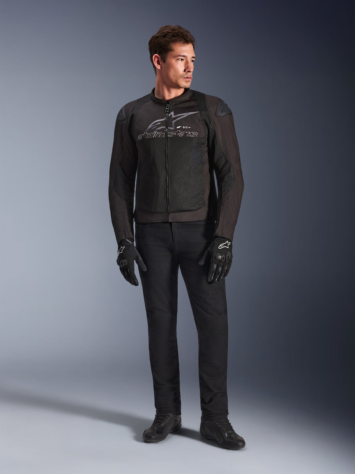 Modello indossa la giacca Alpinestars SMX Air, giacca da moto Black Anthracite con pannelli in rete ventilata e logo grigio sul petto, abbinata a pantaloni neri e guanti protettivi, ambientazione in studio con sfondo sfumato