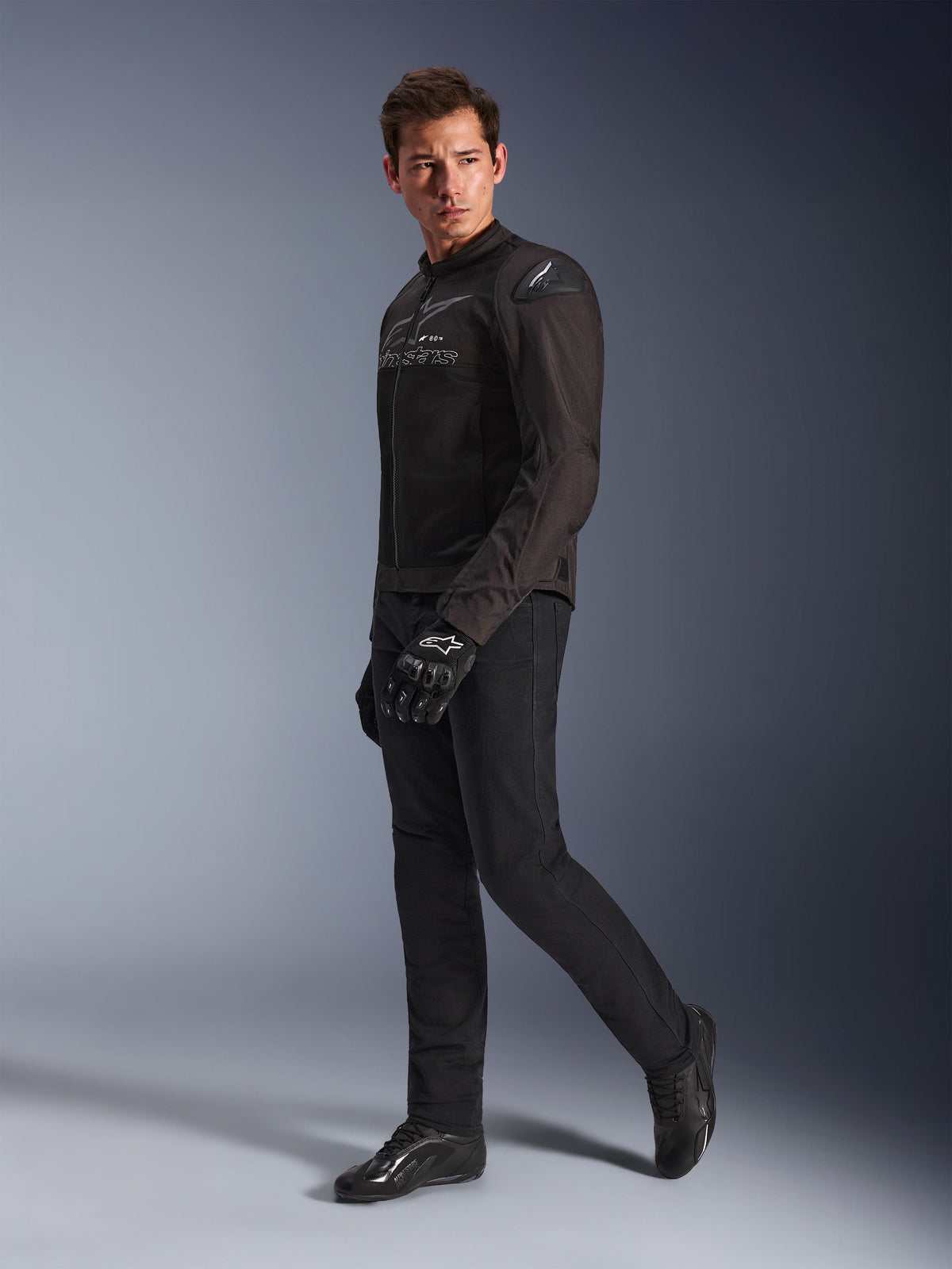 Giacca Alpinestars SMX Air in Black Anthracite, indossata da un modello con pantaloni da moto, guanti e scarpe neri coordinati, inquadratura a figura intera in studio, abbigliamento tecnico sportivo da moto con mesh tecnica e slider sulle spalle
