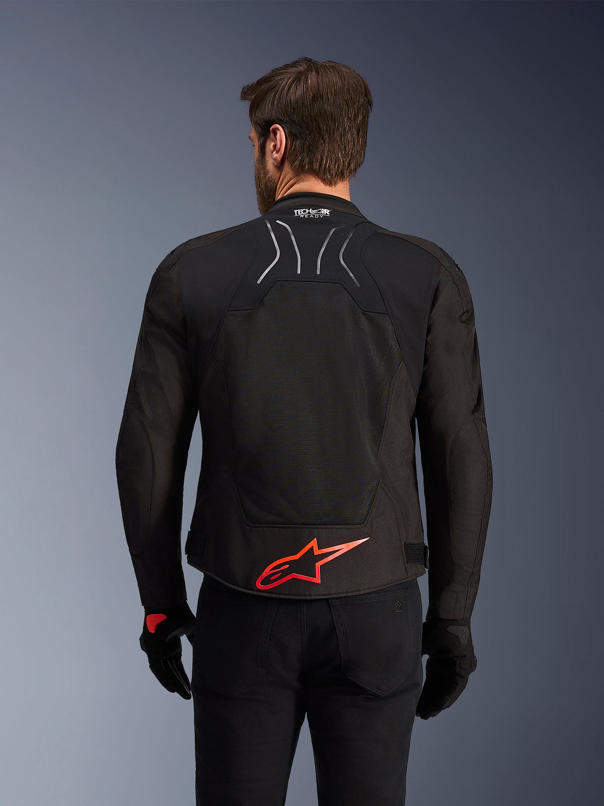 Giacca Alpinestars SMX Air, vista posteriore, nera con logo Alpinestars rosso acceso sulla parte inferiore della schiena, pannello in rete tecnica ad alta ventilazione e marchio Tech-Air Ready, indossata da un pilota con pantaloni e guanti neri