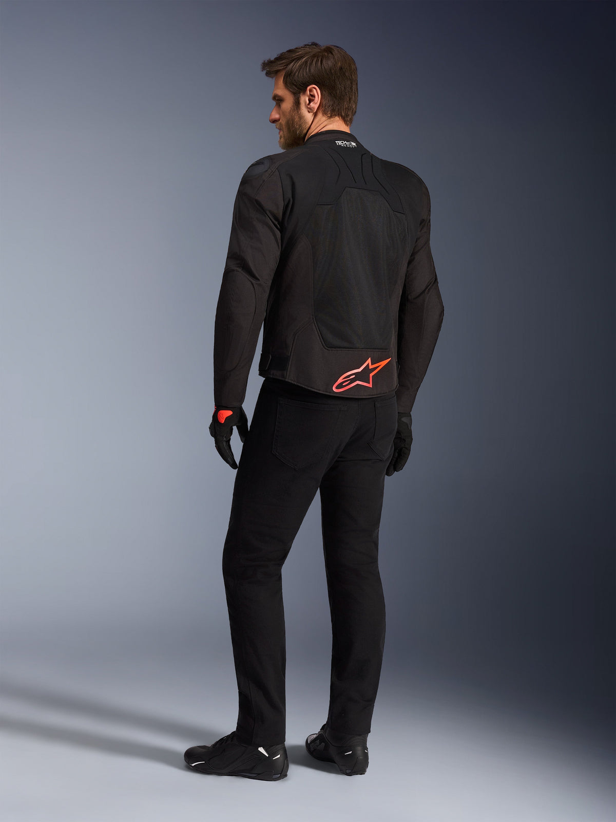Vista posteriore di un modello che indossa la giacca Alpinestars SMX Air in nero, con logo Alpinestars rosso acceso sulla parte bassa della schiena, marchio 'Tech-Air Ready' sul colletto e pannelli in rete tecnica per la ventilazione, abbinata a pantaloni e guanti da moto neri.