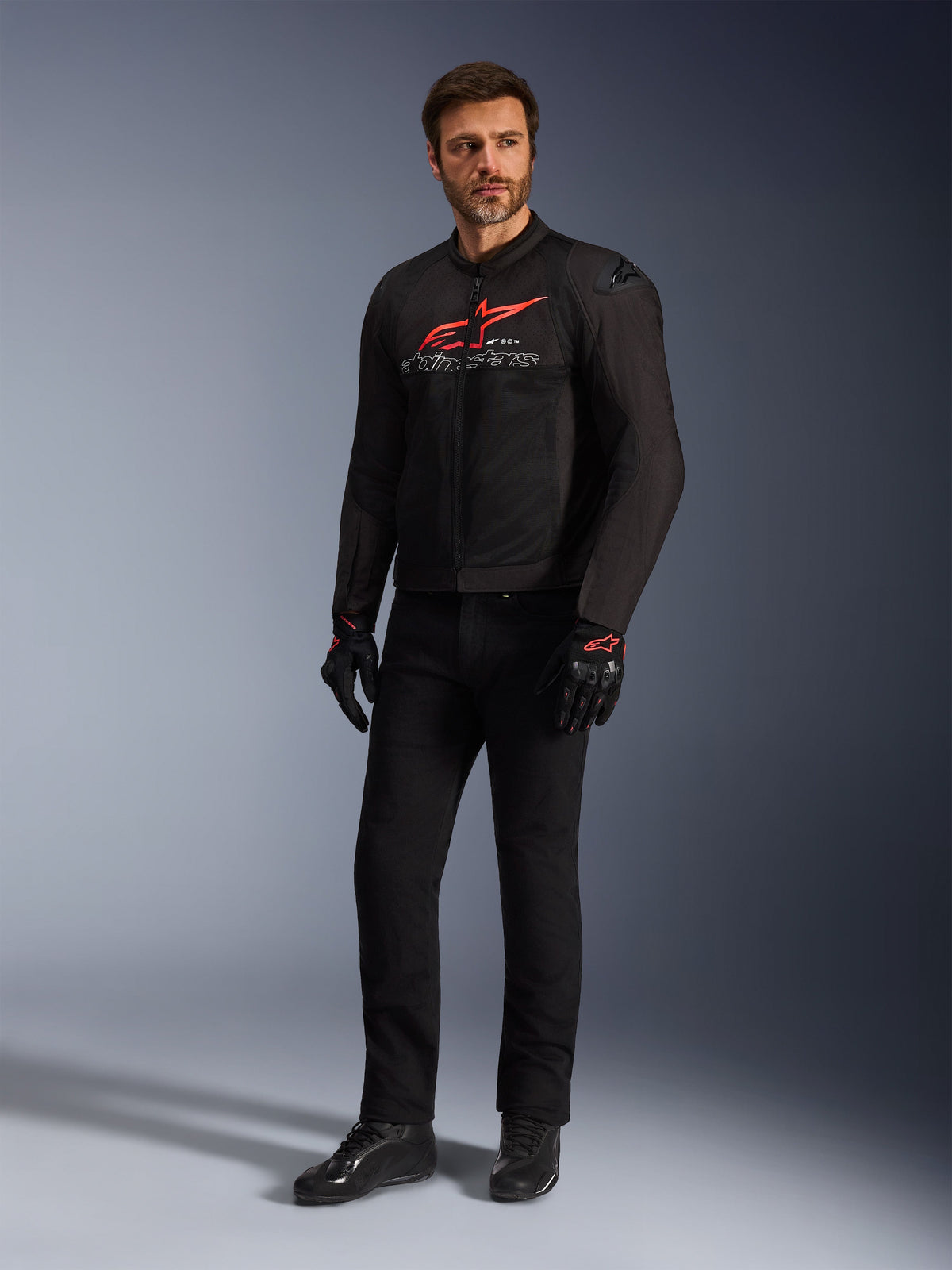 Modello uomo che indossa la giacca Alpinestars SMX Air in Nero Rosso Brillante Bianco, abbinata a pantaloni tecnici neri, guanti neri e rossi e scarpe da guida nere, con un evidente logo rosso sul petto e branding bianco, scatto in studio a figura intera per abbigliamento sportivo da motociclismo