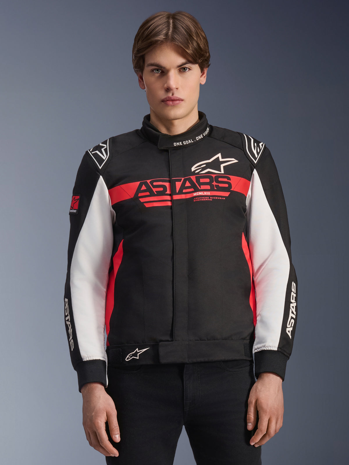 Giacca Alpinestars Monza Sport, giacca da moto in tessuto, nero, rosso vivace e bianco, con vistoso logo ASTARS sul petto, scritta 'ONE GOAL. ONE VISION.' sul colletto, pannelli bianchi sulle maniche e loghi a stella, indossata da un modello su uno sfondo sfumato