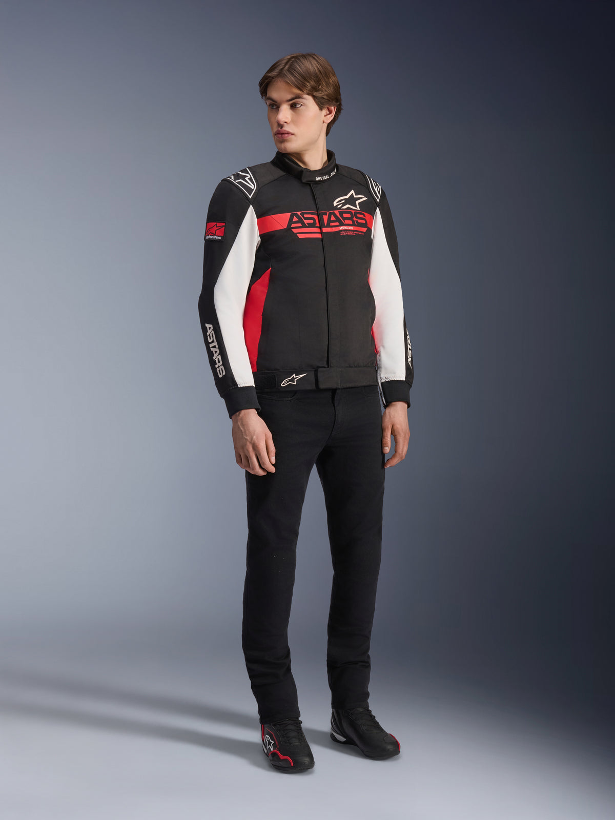 Giacca Alpinestars Monza Sport, nero, rosso acceso e bianco, giacca da moto in tessuto, design ispirato alle corse con marchio ASTARS su petto e maniche, indossata da un modello con pantaloni neri e scarpe da moto, abbigliamento da guida casual sportivo