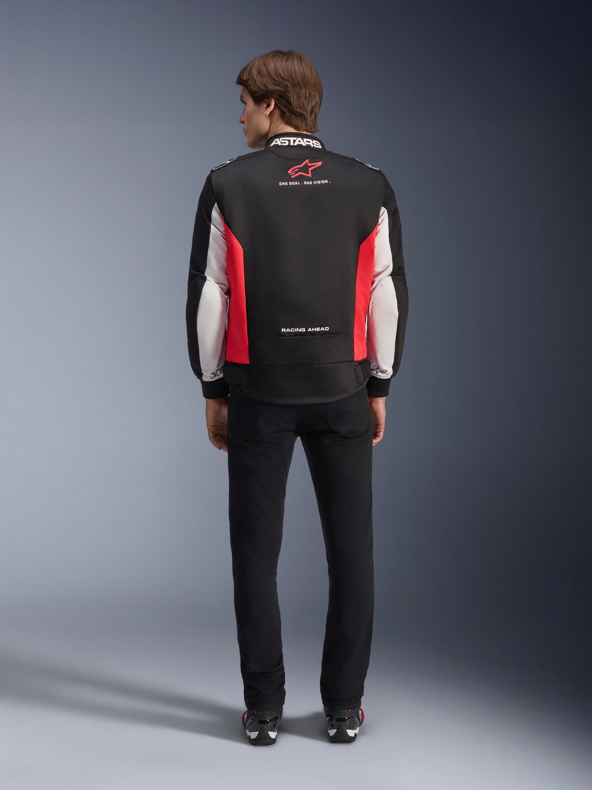Giacca Alpinestars Monza Sport, giacca da moto in tessuto nero, rosso acceso e bianco, vista posteriore su un modello, con logo a stella rossa e marchio 'ASTARS' sul colletto con slogan, indossata con pantaloni neri e stivali da moto.