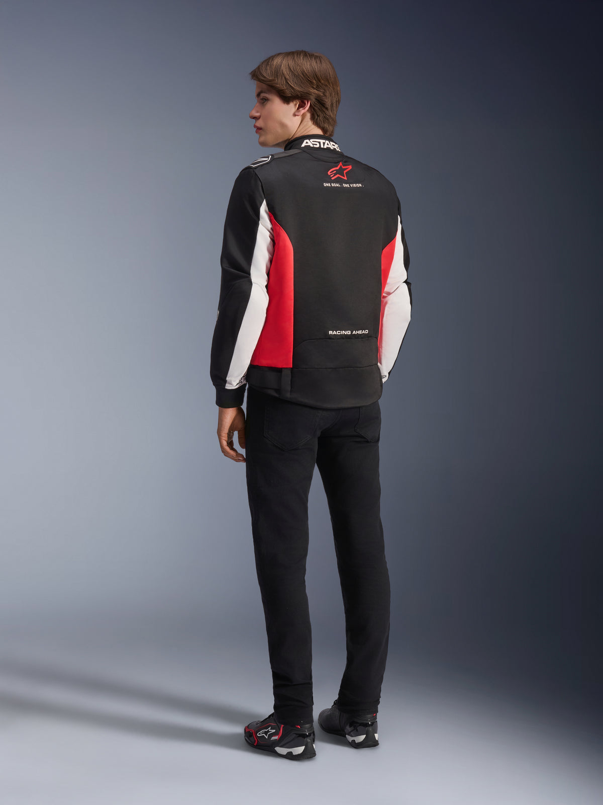 Giacca Alpinestars Monza Sport, giacca da moto in tessuto, nero, rosso brillante e bianco, vista posteriore del modello con pannelli laterali rossi e dettagli bianchi sulle maniche, logo Alpinestars e scritta 'ONE GOAL. ONE VISION.', abbinata a pantaloni neri e scarpe da guida per un look d'ispirazione sportiva.