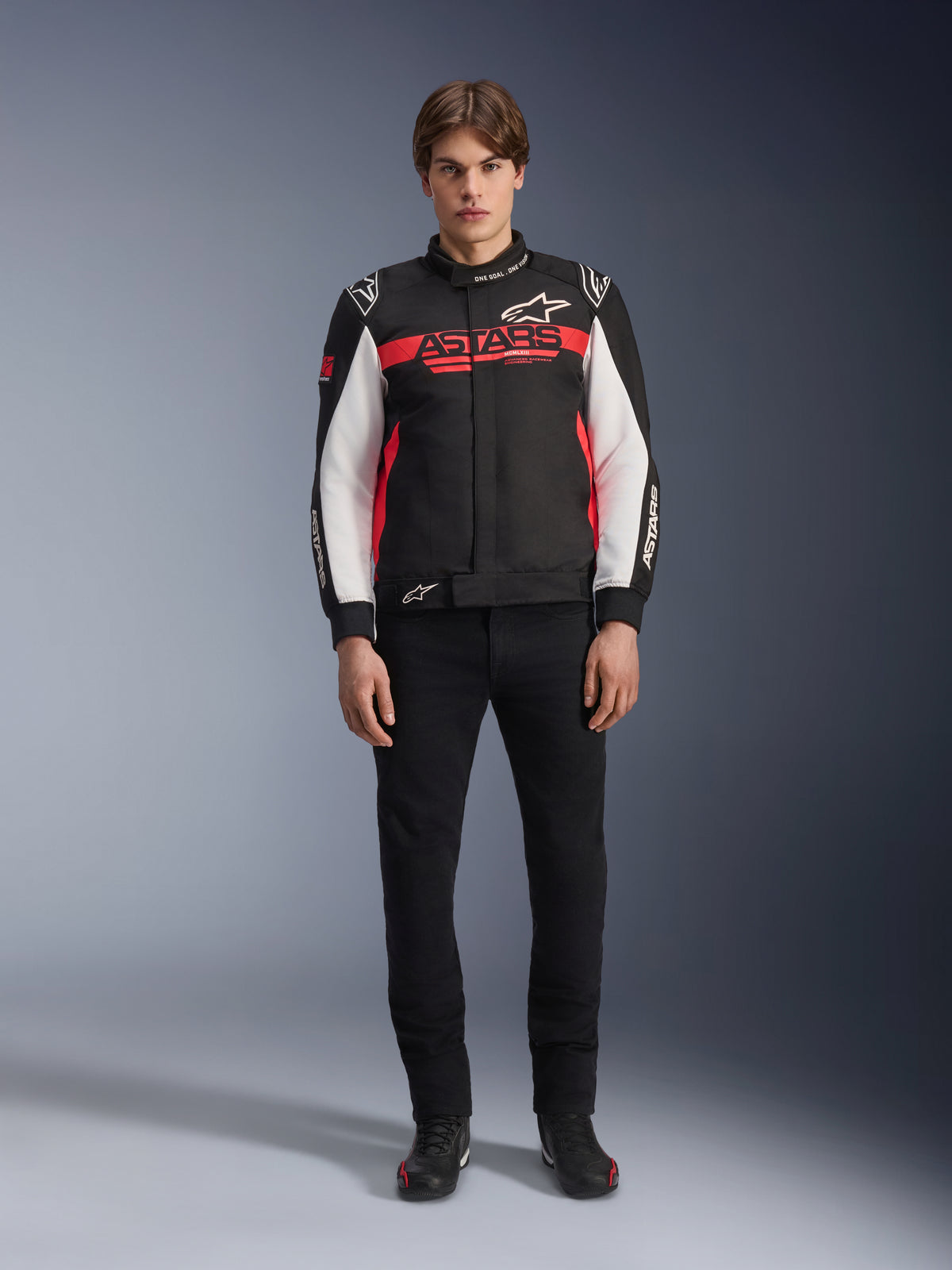 Modello che indossa la giacca Alpinestars Monza Sport in nero, rosso acceso e bianco, con un grande logo 'ASTARS' rosso sul petto e pannelli bianchi sulle maniche, abbinata a pantaloni neri e stivali da moto neri.