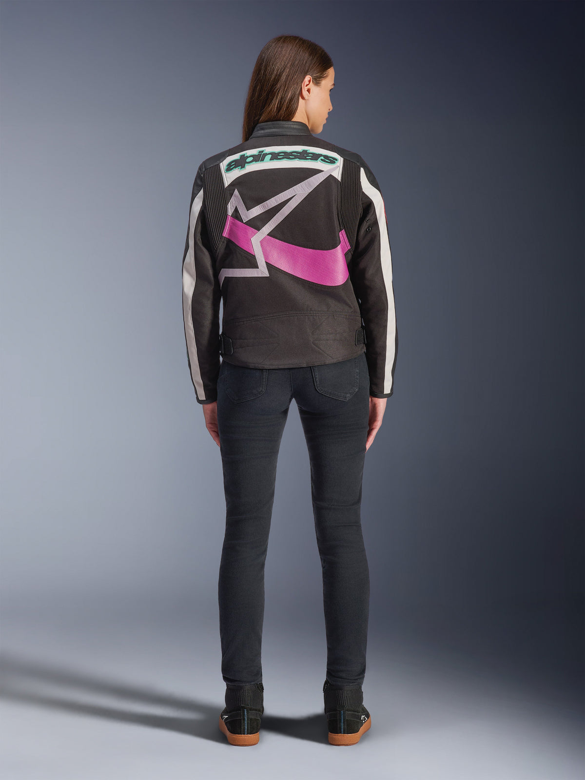 Vista posteriore di una donna che indossa la giacca Alpinestars Idles, nera con strisce bianco sporco, caratterizzata da un grande logo Alpinestars a stella viola e bianco e una scritta verde sul retro, abbinata a jeans slim-fit scuri, abbigliamento da moto urbano d'ispirazione vintage