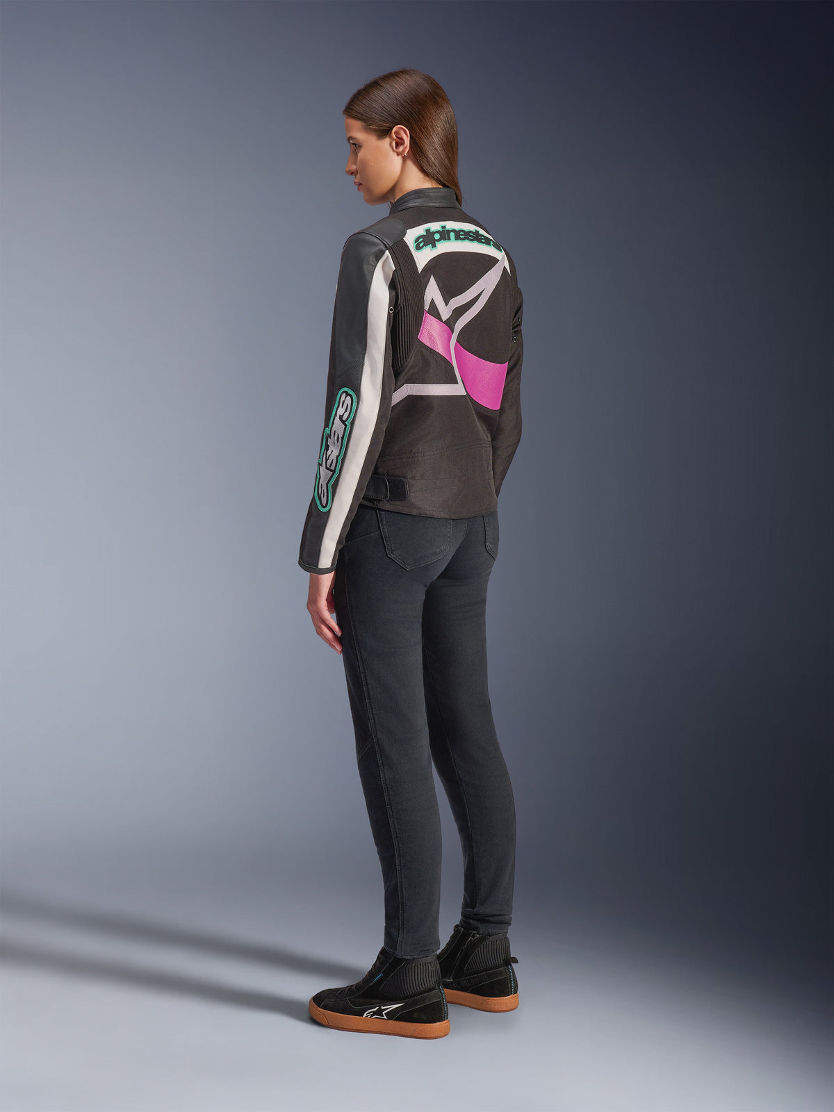 Giacca Alpinestars Idles da donna, giacca da moto, nero, bianco sporco e viola, vista dal retro su modella, con un grande logo Alpinestars avvolgente e design d'ispirazione racing vintage, abbinata a jeans scuri e scarpe nere.