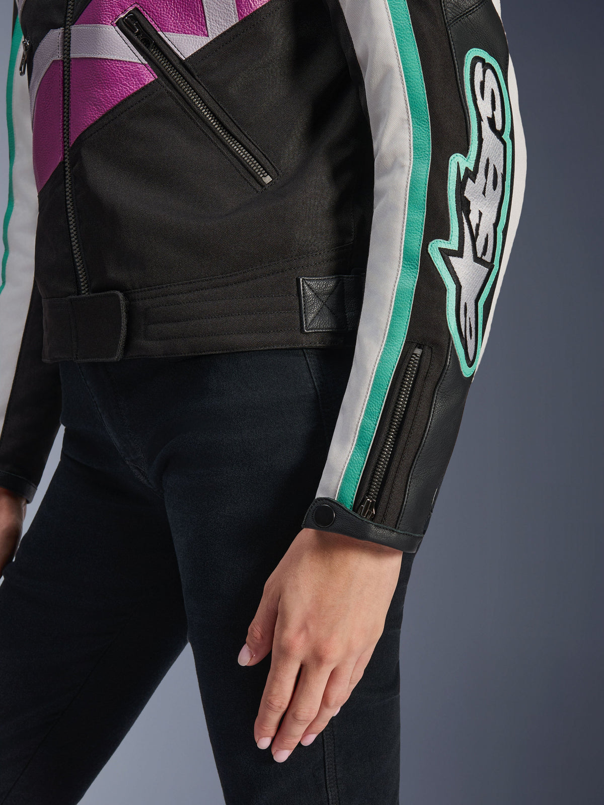 Giacca da donna Alpinestars Idles, vista ravvicinata della manica e del busto laterale, nera con dettagli viola, bianchi e verde acqua, con un audace logo Alpinestars sul braccio, polsino con zip e bottone a pressione e cinturino di regolazione in vita, abbigliamento da moto d'ispirazione vintage.