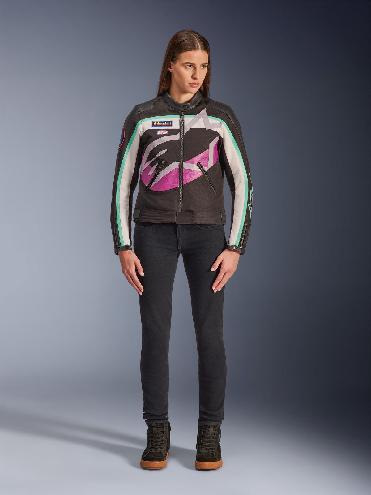 Donna che indossa la giacca Alpinestars Idles Donna, nero, bianco sporco e viola, design di ispirazione racing vintage con grande logo avvolgente e accenti verde acqua, abbinata a jeans neri e sneakers da moto, abbigliamento motociclistico urbano