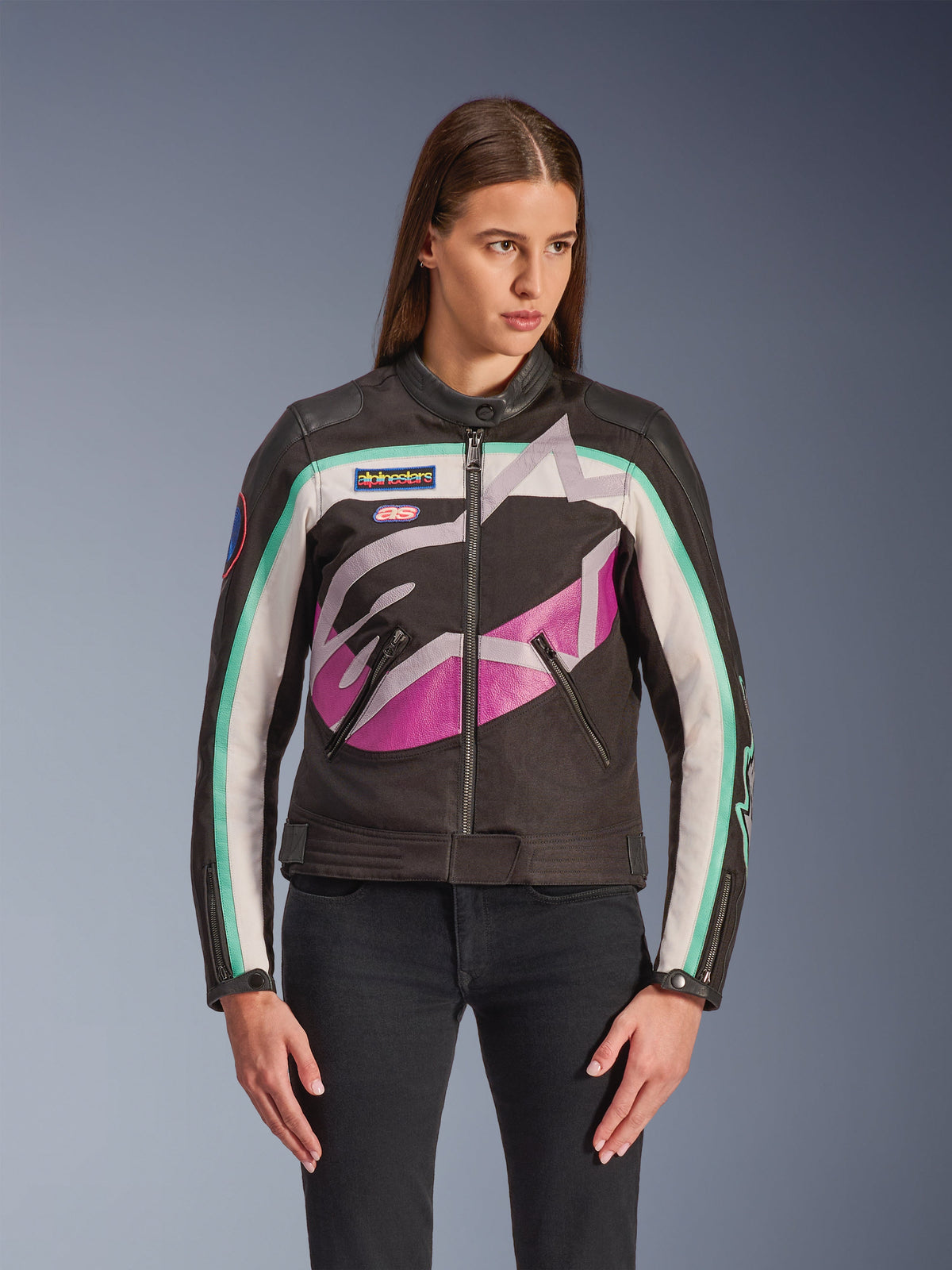 Giacca da donna Alpinestars Idles, giacca da moto, nera con design color block bianco, viola e teal, stile d'ispirazione vintage racing con grande logo astratto sul petto, patch del marchio e zip frontale, indossata da una modella con jeans neri