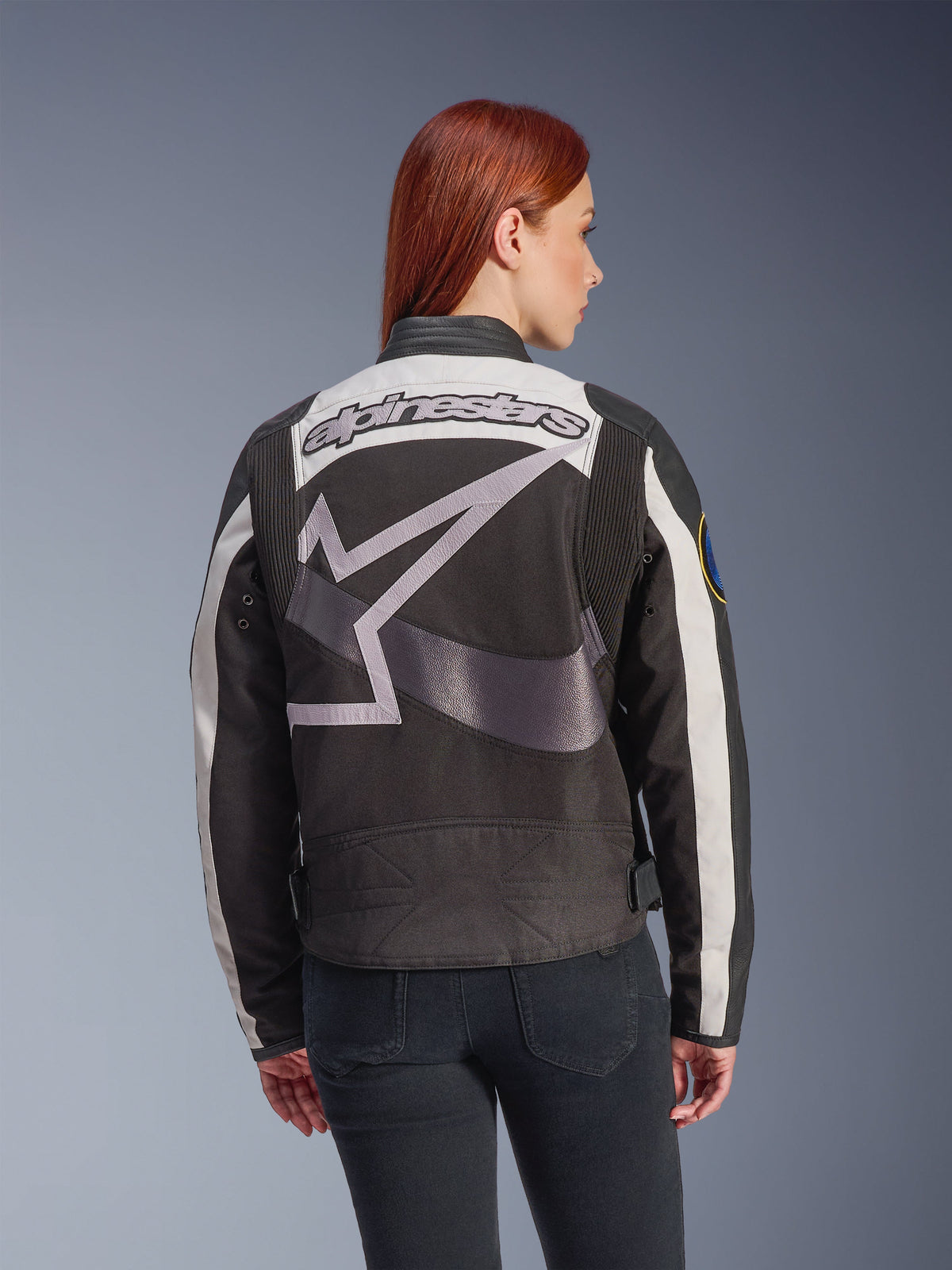 Giacca da donna Alpinestars Idles, nero e bianco sporco, vista posteriore con grande logo a stella grigio e scritta Alpinestars, indossata da una modella con i capelli rossi, giacca da moto urbana d'ispirazione vintage con pannelli a fisarmonica.