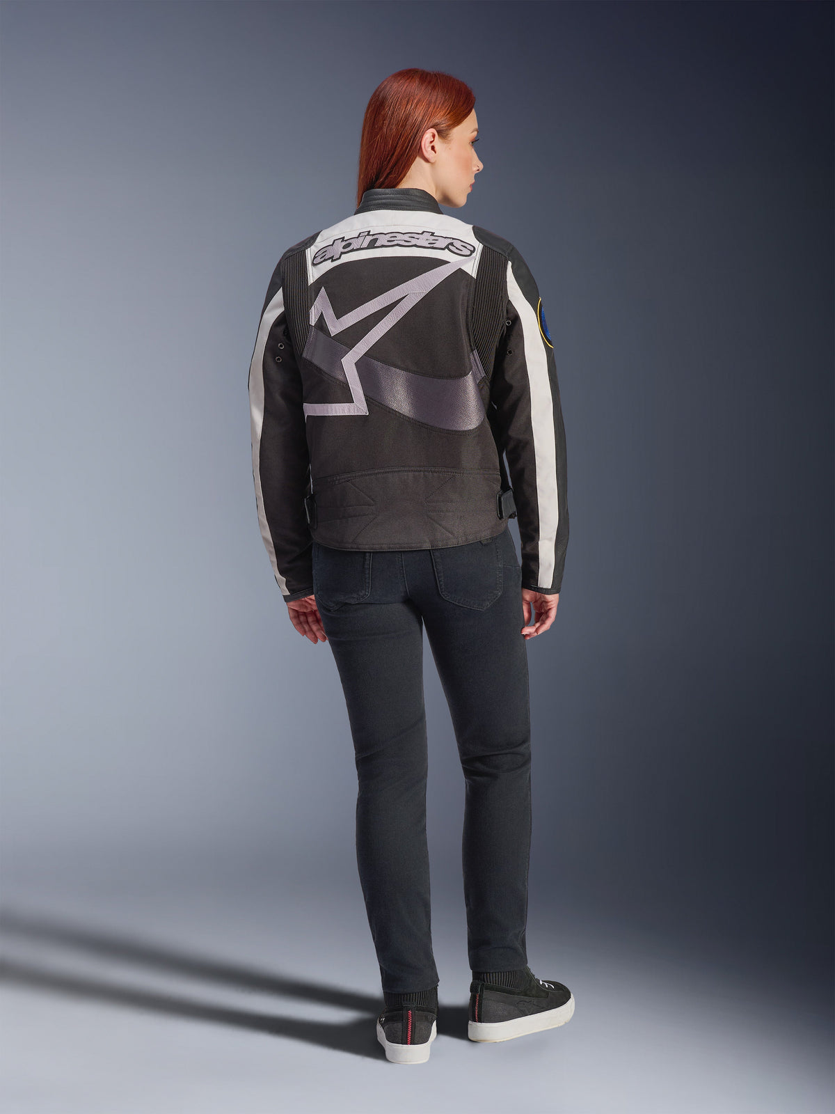 Giacca da donna Alpinestars Idles, giacca da moto urban, nero e bianco sporco, vista posteriore con un grande logo e scritta Alpinestars grigi, indossata con jeans neri slim-fit e sneakers, design d'ispirazione vintage per la guida urbana