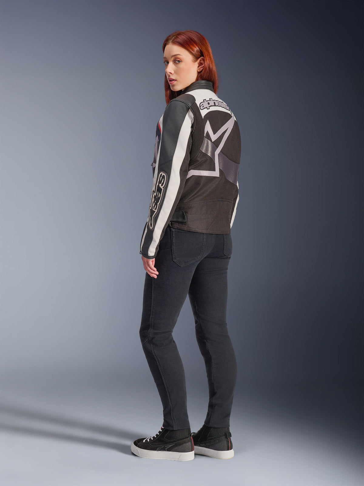 Donna che indossa la giacca Alpinestars Idles, vista posteriore, Off White, Rosso Fiamma e Nero, con un grande logo a stella sul retro e scritta Alpinestars sulla manica, abbinata a pantaloni neri slim-fit e sneakers, abbigliamento moto urbano da donna
