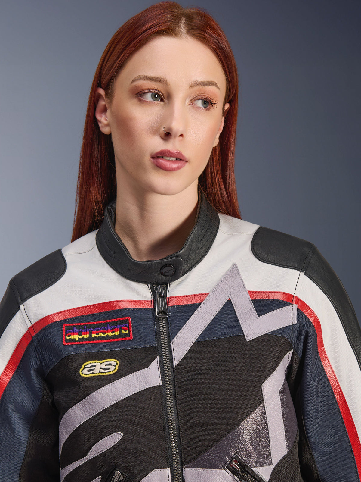 Giacca da donna Alpinestars Idles, primo piano di una modella con capelli rossi, colorazione Off White, Flame Red e Black, design racing d'ispirazione vintage con patch logo e grafiche a stella geometriche grigie, abbigliamento tecnico da moto urbano.