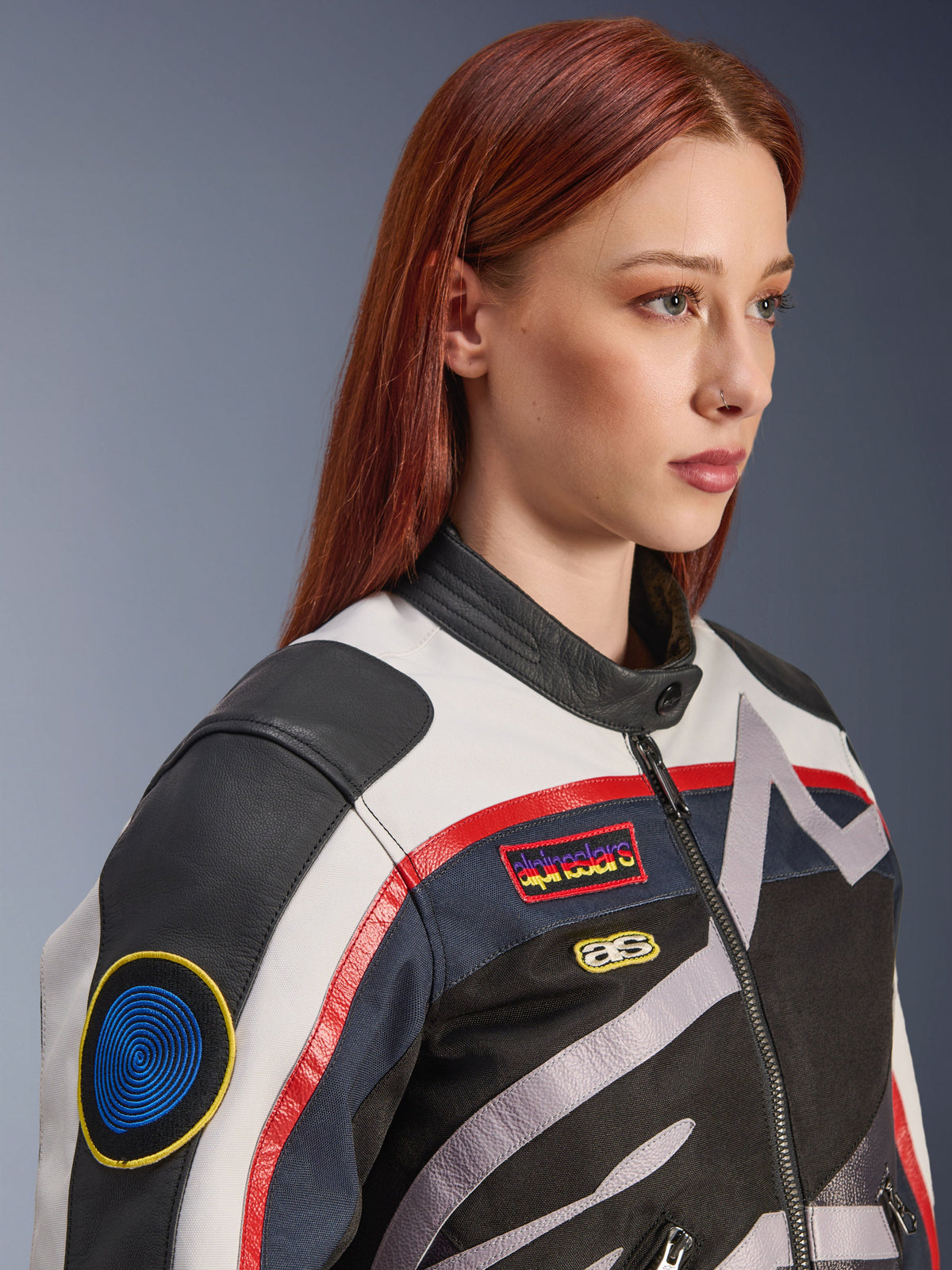 Giacca da donna Alpinestars Idles, bianco panna, rosso fiamma e nero, con toppe da corsa d'ispirazione vintage e logo avvolgente, struttura in tessuto e pelle, stile motociclistico urbano indossata da una modella