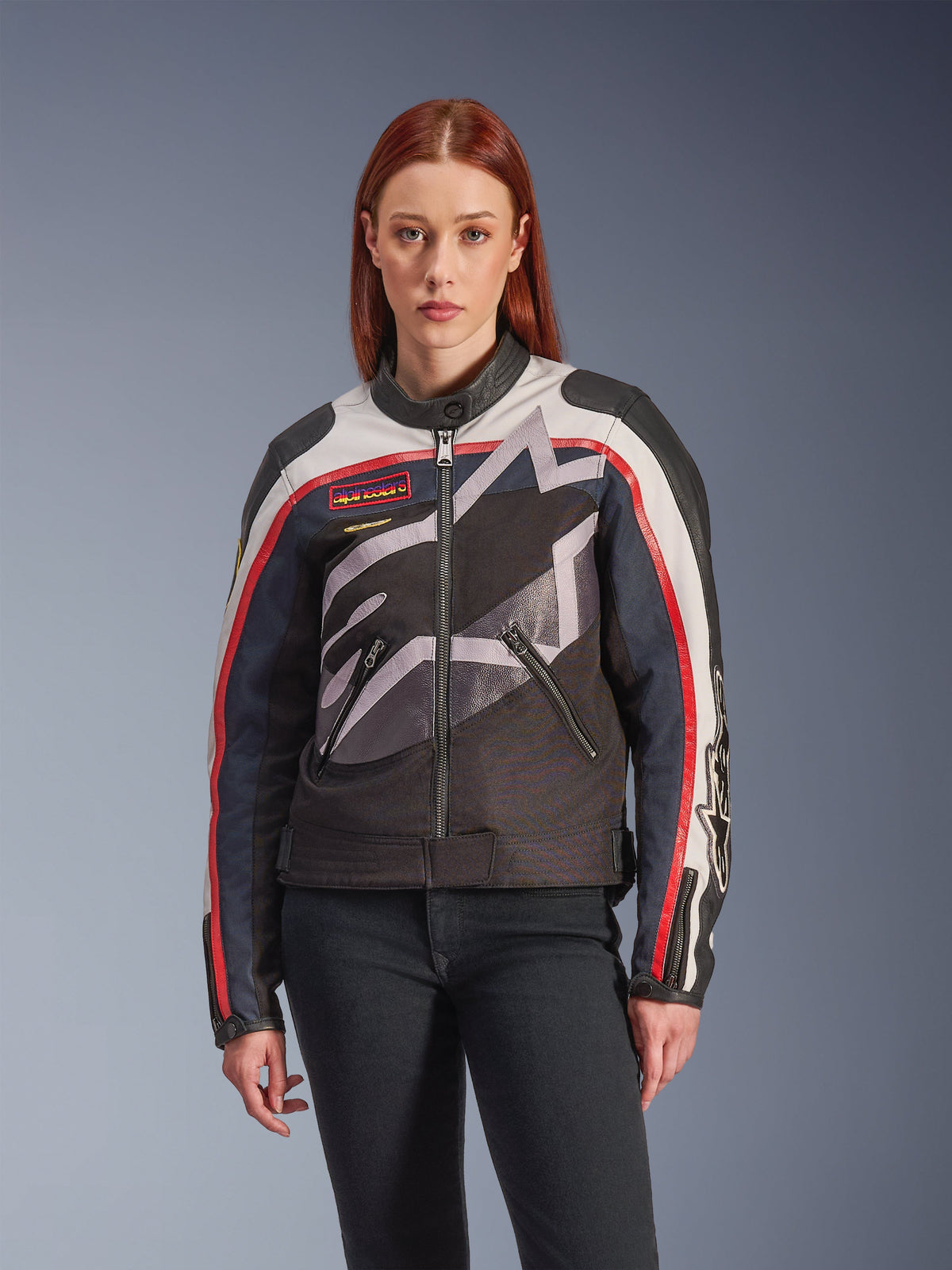 Giacca Alpinestars Idles, giacca da moto da donna, bianco sporco, rosso fiamma e nero, con un grande logo a stella stilizzato, patch sul petto d'ispirazione vintage, collo alto stile biker, tasche con zip e maniche a strisce, indossata da una modella con jeans scuri.