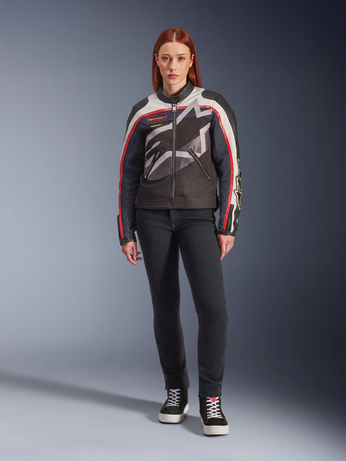 Modella che indossa la giacca Alpinestars Idles, giacca da moto da donna, Off White Flame Red e nero, caratterizzata da un grande logo a stella avvolgente e branding d'ispirazione vintage, abbinata a pantaloni da guida scuri slim-fit e sneaker alte nere, abbigliamento da moto urban contemporary