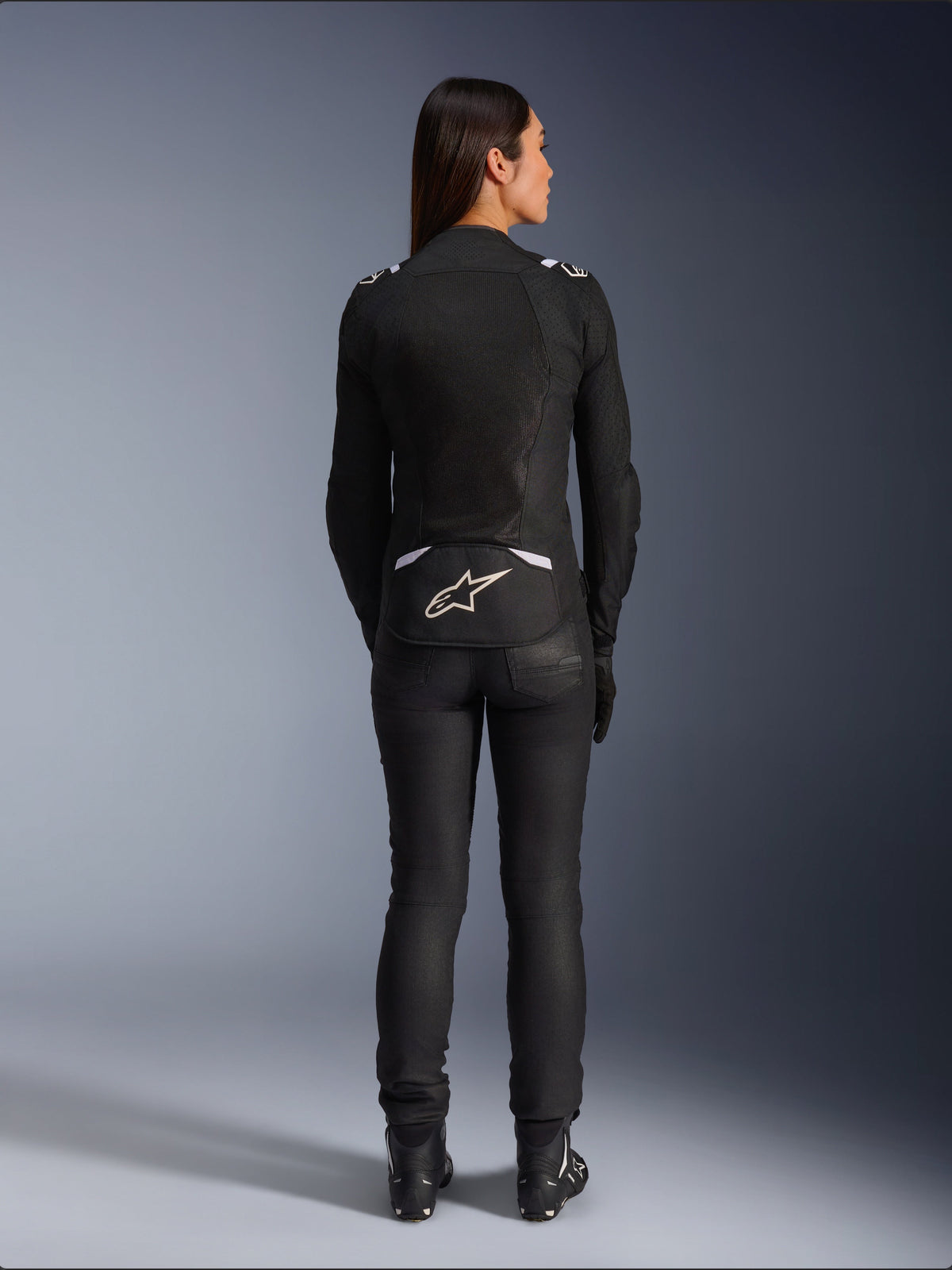 Giacca Alpinestars Stella T-SPS Air v2, giacca da moto in tessuto da donna, nero e bianco, vista posteriore con pannelli di ventilazione in mesh e logo bianco sulla parte bassa della schiena, indossata con pantaloni da guida e stivali neri, abbigliamento sportivo urbano
