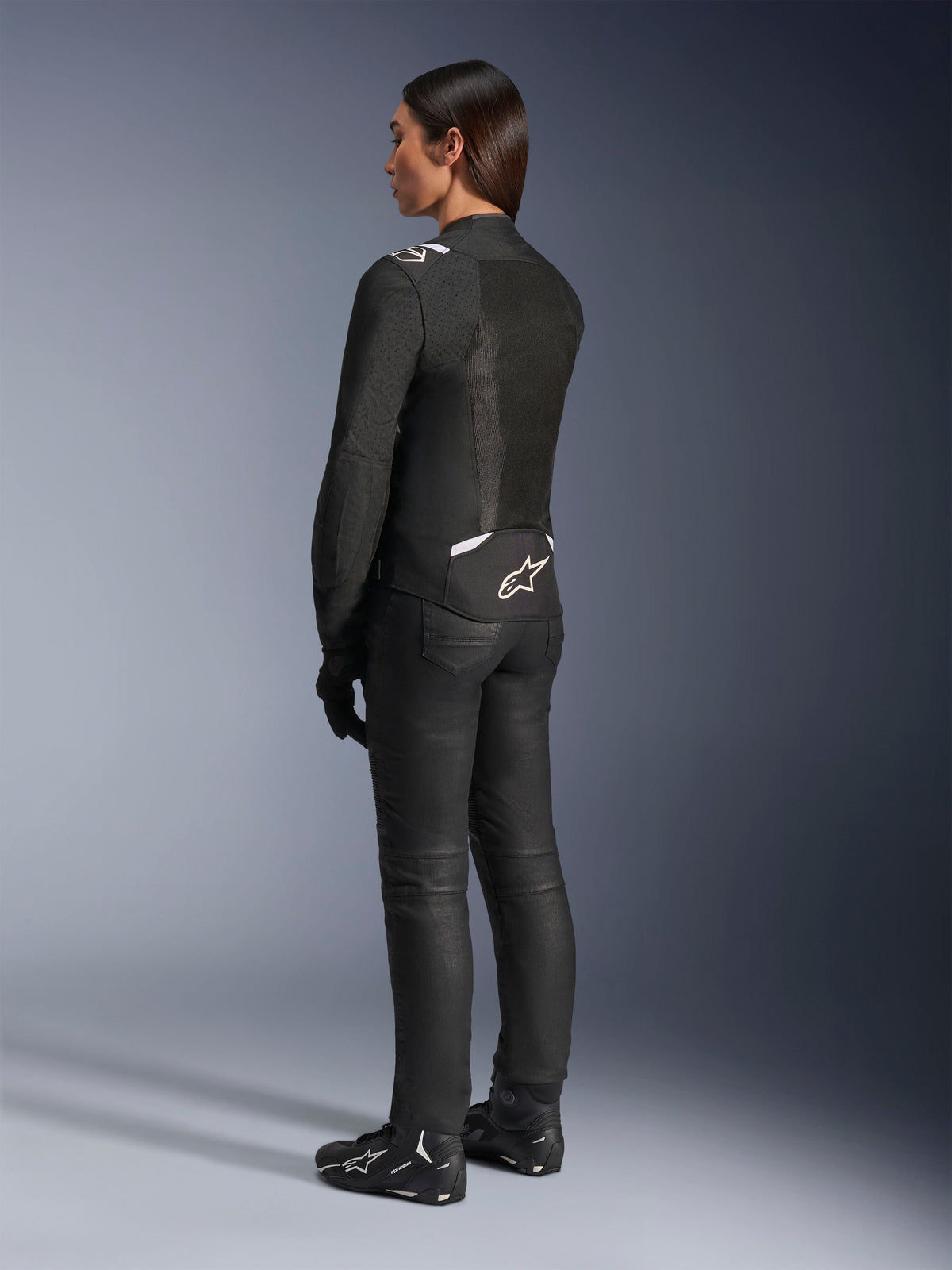 Giacca Alpinestars Stella T-Sps Air V2, nero e bianco, vista posteriore con pannelli in mesh e logo sulla parte bassa della schiena, abbinata a pantaloni da guida neri e scarpe da moto, abbigliamento tecnico sportivo da strada per donna