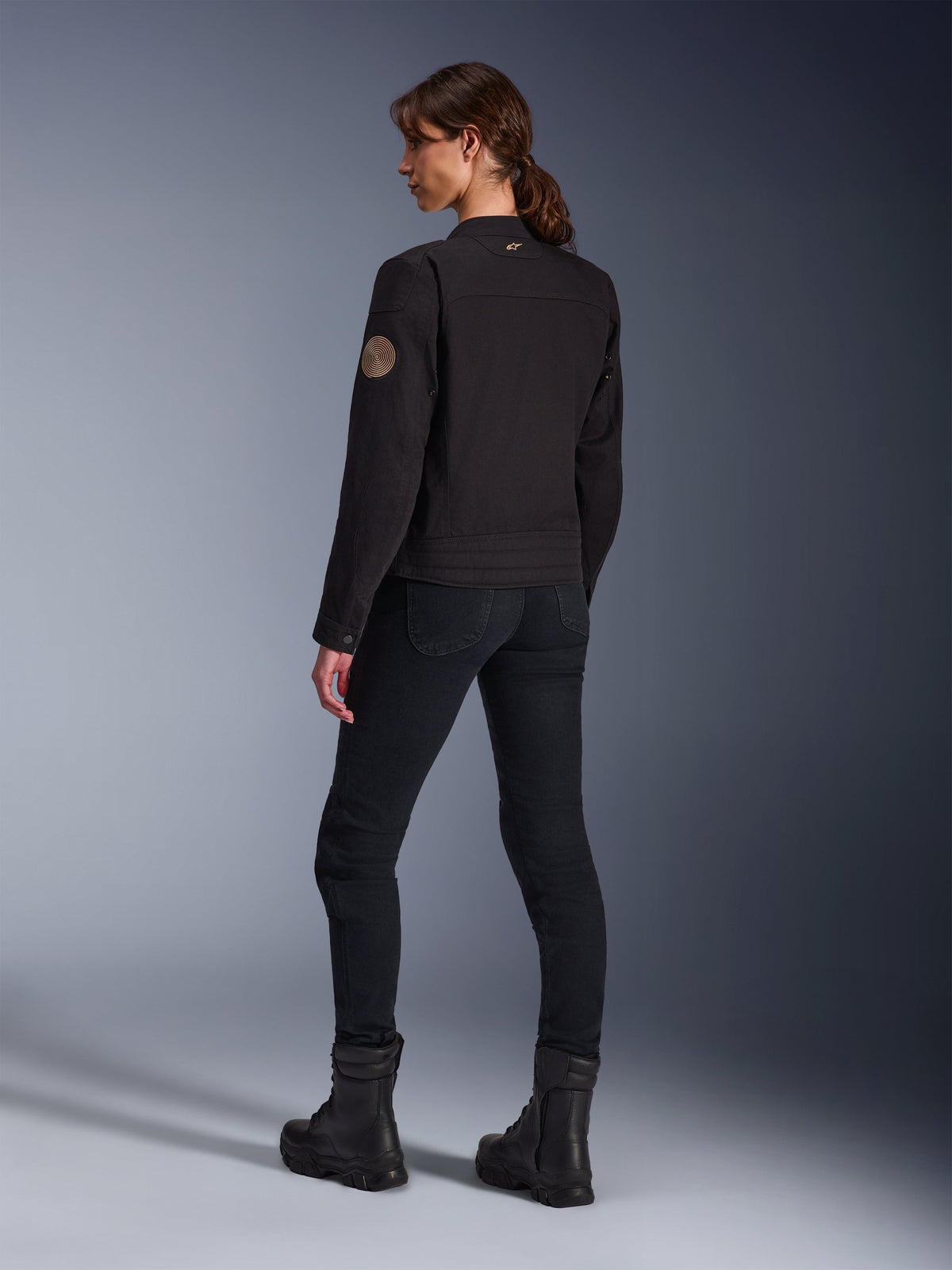Modella che indossa la giacca Alpinestars Stella Grace in nero, vista posteriore che mette in risalto la toppa vintage Oscar a spirale sul braccio e il piccolo logo a stella sul colletto, abbinata a pantaloni da moto e stivali neri per la guida urbana