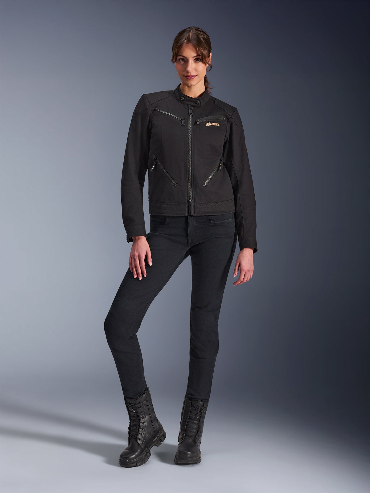 Donna che indossa la giacca Alpinestars Stella Grace in nero, giacca da moto urbana dall'estetica workwear, con tasche anteriori con zip e logo sul petto, abbinata a pantaloni neri slim-fit e stivali neri, abbigliamento da moto contemporaneo da donna.