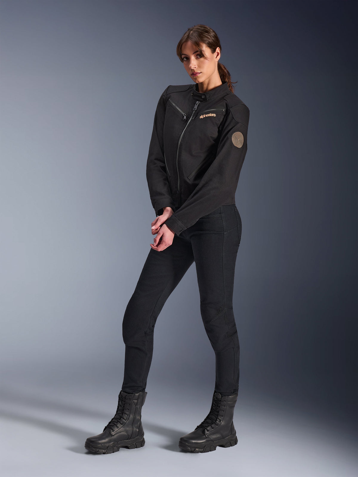 Modella che indossa la Giacca Alpinestars Stella Grace, giacca da moto urbana nera, abbinata a pantaloni da guida e stivali neri, design d'ispirazione workwear con logo ricamato color bronzo sul petto e patch circolare a spirale sulla manica
