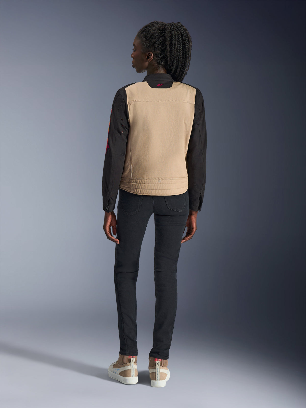 Donna che indossa la giacca Alpinestars Stella Grace, Aluminum Black con busto tan e maniche nere, vista posteriore, abbinata a pantaloni neri slim-fit e sneakers casual, abbigliamento da moto urbano da donna