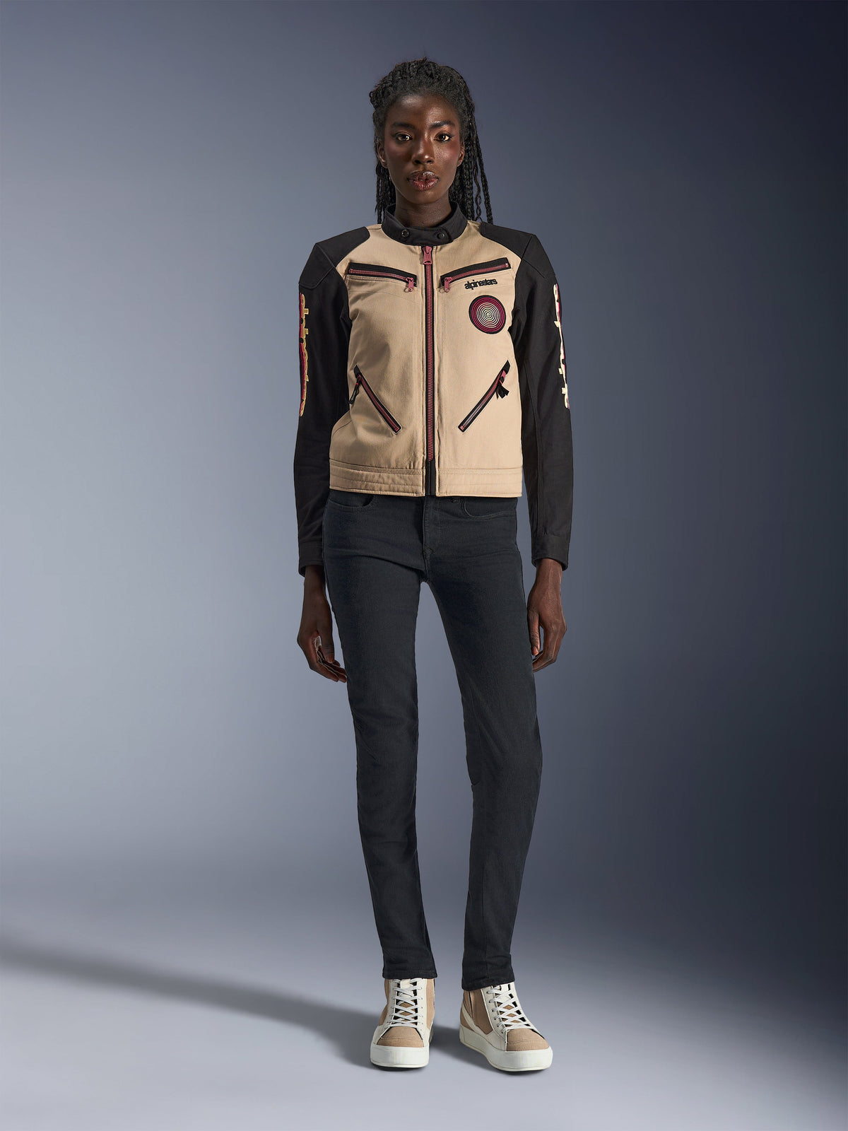 Giacca Alpinestars Stella Grace da donna, giacca da moto urban, costruzione in tessuto color sabbia e nero, con cerniere anteriori e patch Oscar a spirale ricamata, indossata con pantaloni neri e sneaker alte, abbigliamento tecnico da moto ispirato al workwear contemporaneo