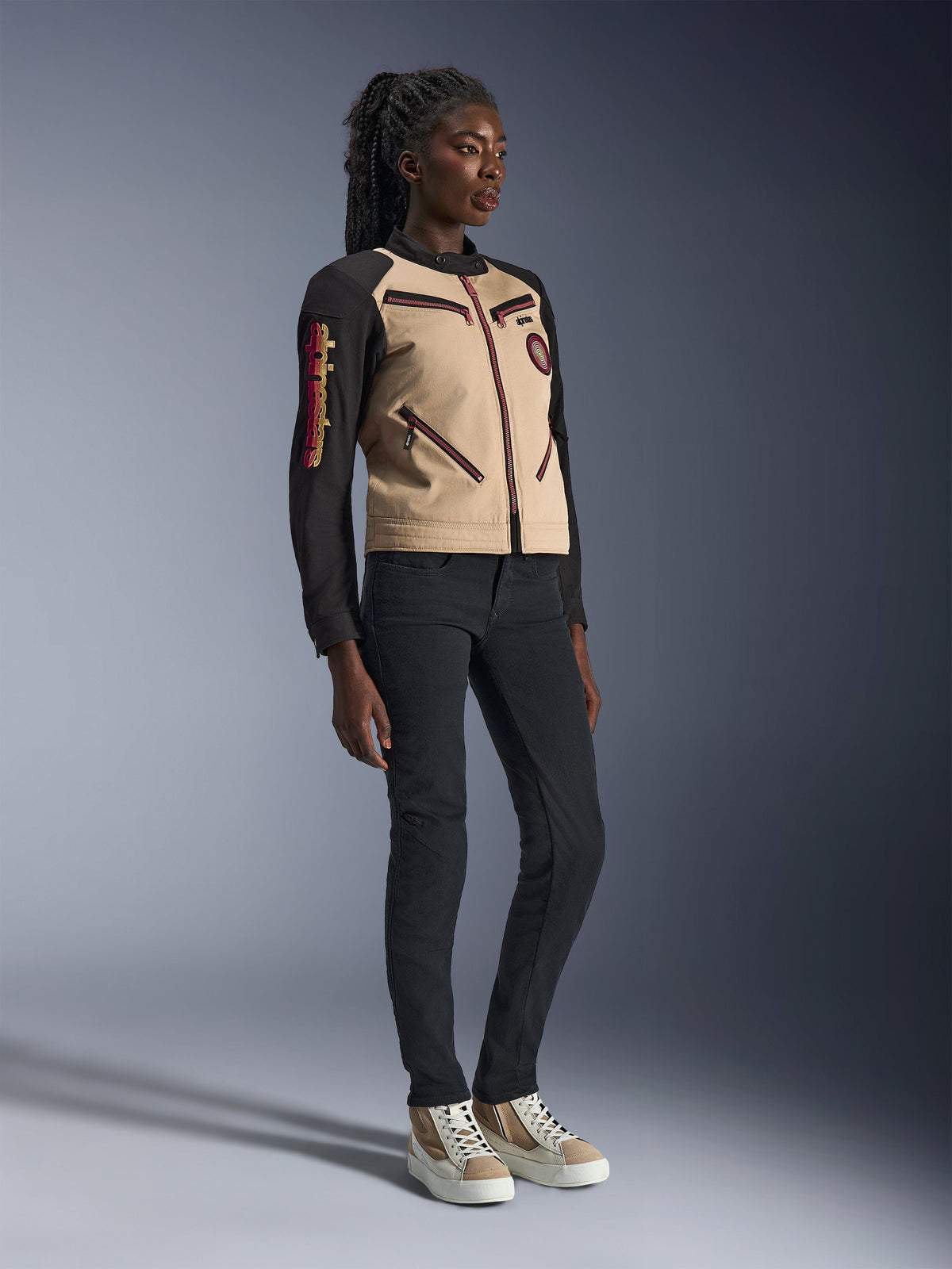Modella che indossa la giacca Alpinestars Stella Grace in Aluminum Black, con busto beige e maniche nere con accenti rossi sulle cerniere, abbinata a pantaloni neri slim-fit e sneakers crema, abbigliamento da moto urban contemporary per donna