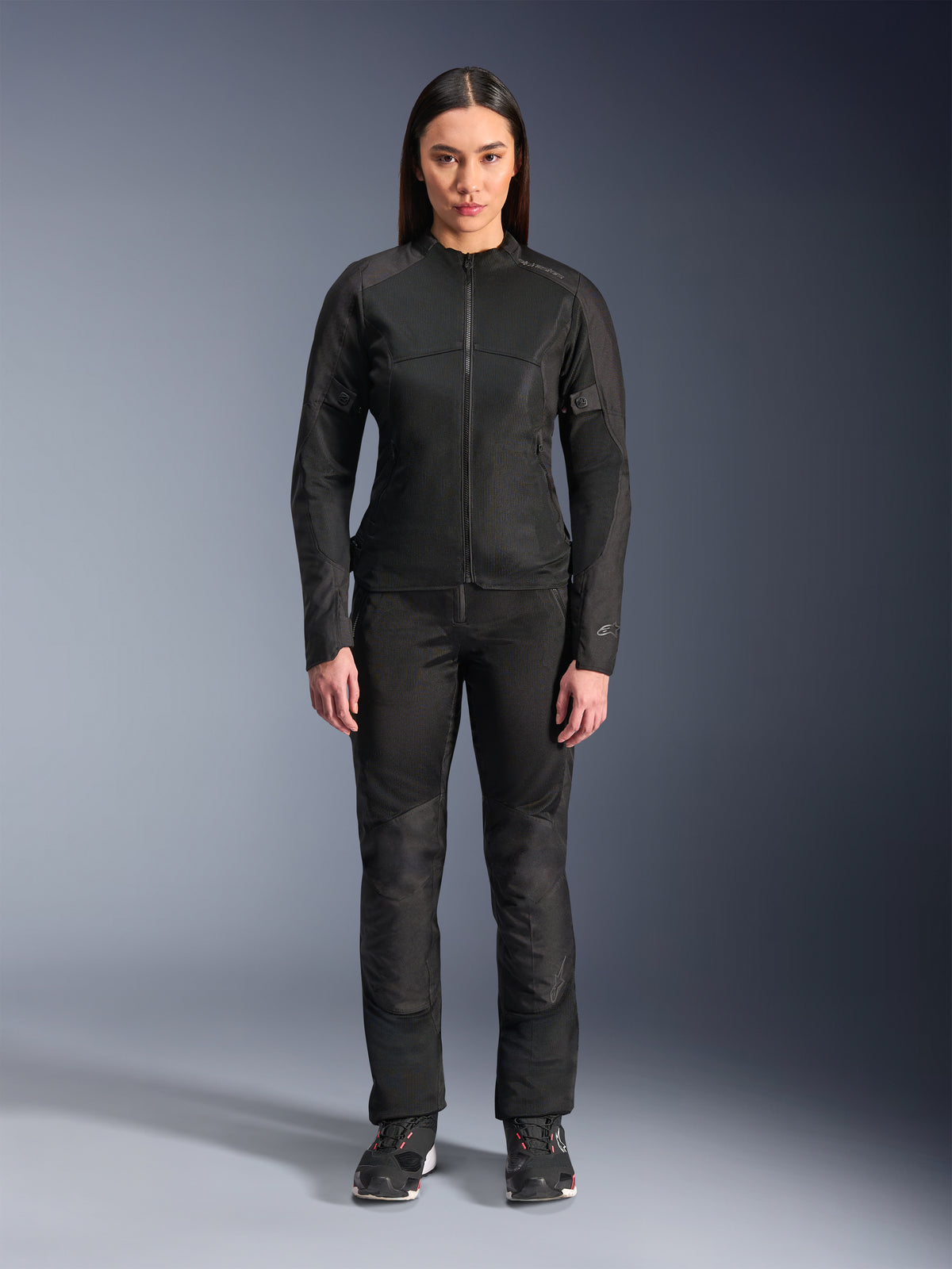 Donna che indossa la giacca Alpinestars Stella C-1 Air in nero, abbinata a pantaloni da moto in tessuto nero coordinati e scarpe da guida, caratterizzata da un design urbano elegante con ampi pannelli in mesh per il flusso d'aria.