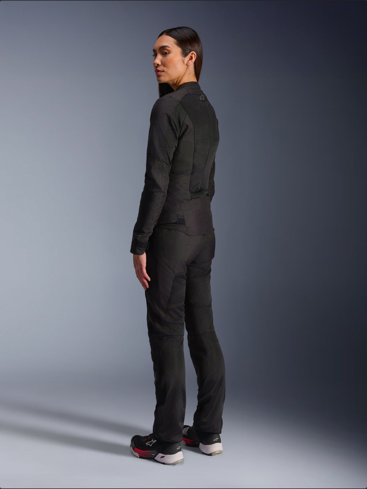 Donna che indossa la giacca Alpinestars Stella C-1 Air in nero, giacca da moto urban in tessuto con pannelli in rete, vista posteriore-laterale che mostra la vestibilità aderente e il logo sulla parte superiore della schiena, indossata con pantaloni neri coordinati e scarpe sportive, abbigliamento da moto contemporaneo.