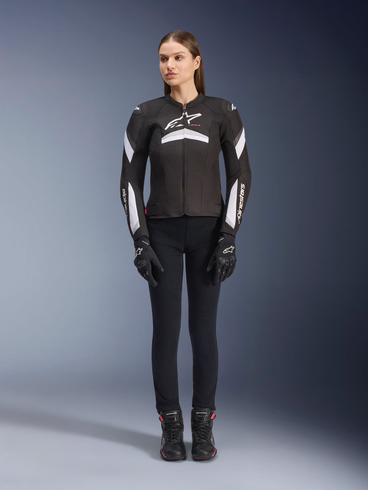 Modella che indossa la giacca Alpinestars Stella T-GP Plus R V4 in nero e bianco, giacca da moto sportiva in tessuto, abbinata a pantaloni da moto, guanti e stivali neri, vista frontale con logo Alpinestars bianco e dettagli sulle maniche.