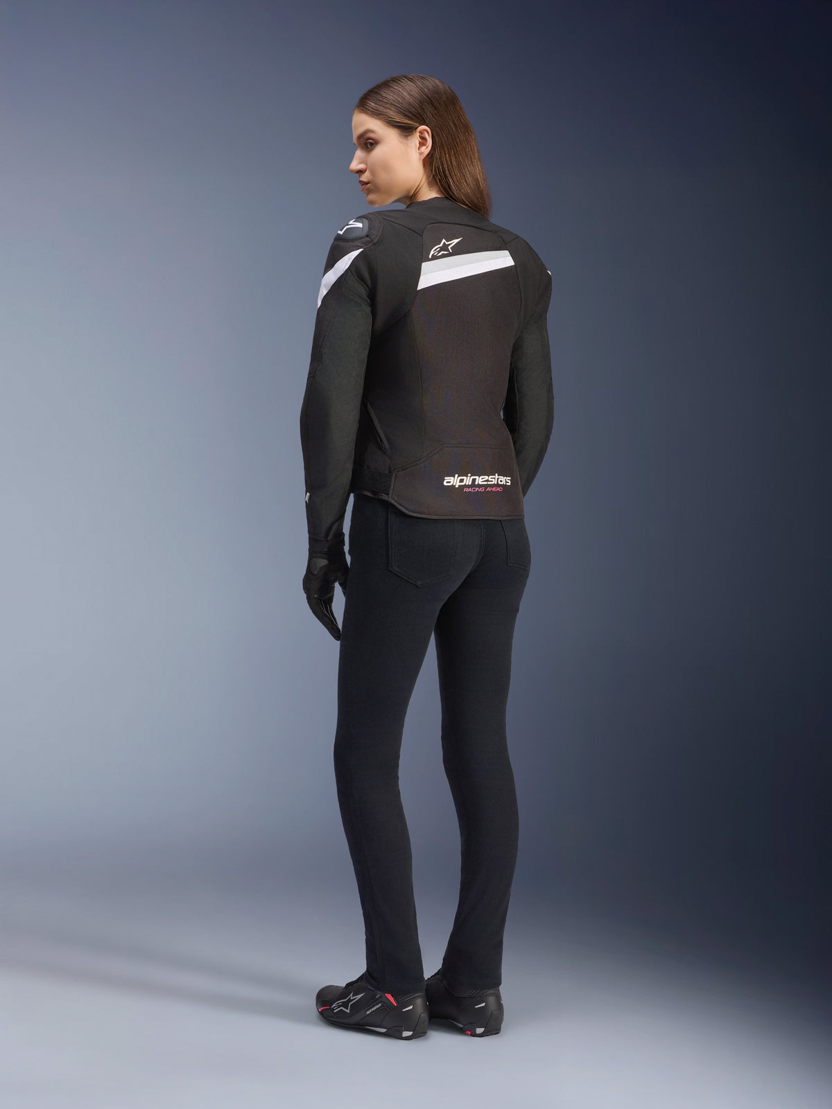 Alpinestars Stella T-GP Plus R V4 Jacket, giacca da moto in tessuto da donna, nero e bianco, vista posteriore su modella, con accenti bianchi sulle spalle e logo Alpinestars, indossata con pantaloni neri e guanti da moto, design sport-fit per guida su strada