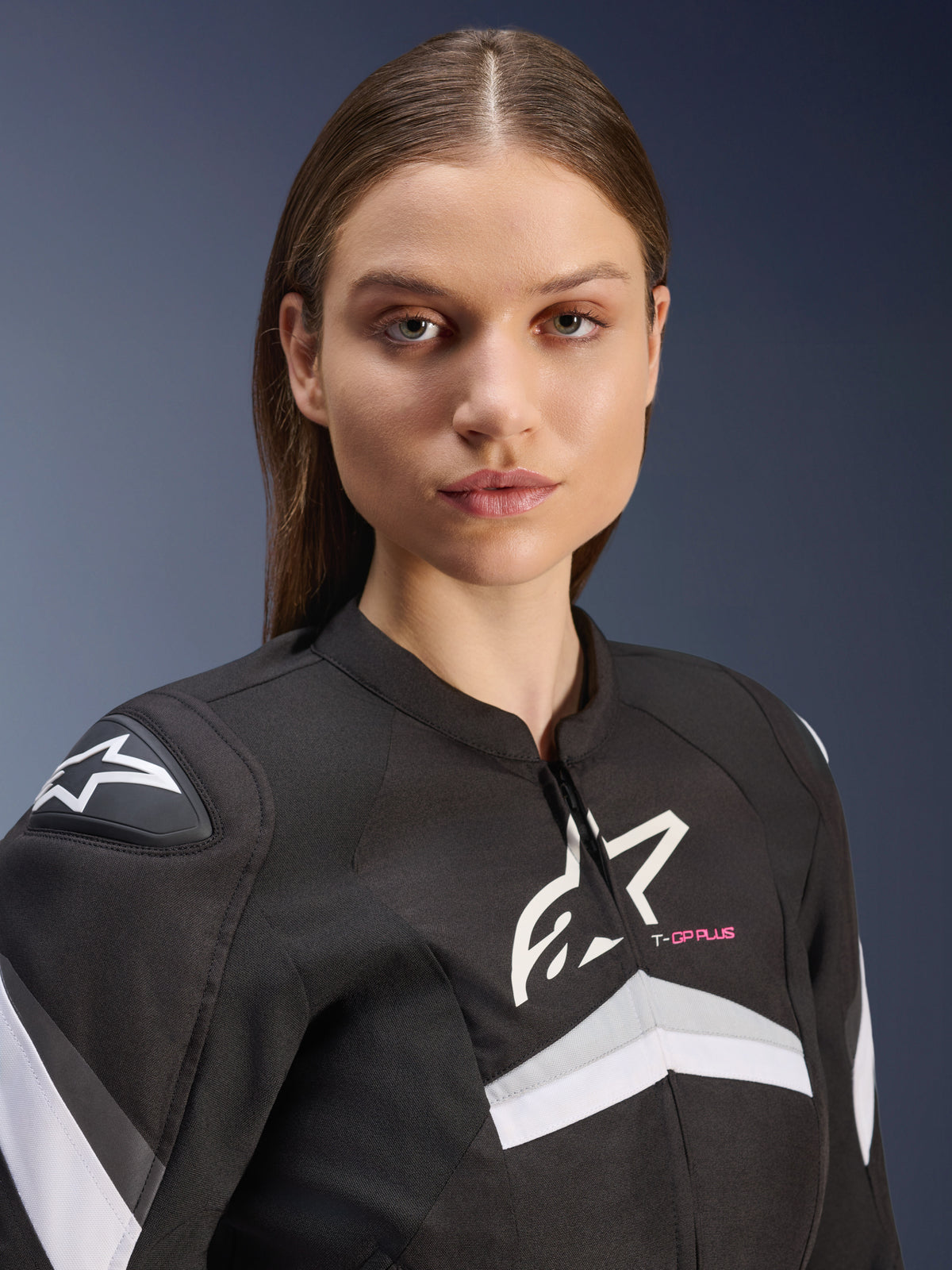 Giacca Alpinestars Stella T-GP Plus R V4, giacca da moto in tessuto da donna, nero e bianco con dettagli grigi e scritta 'T-GP PLUS' in rosa, primo piano di una modella che indossa la giacca con vestibilità sportiva e loghi Astars bianchi