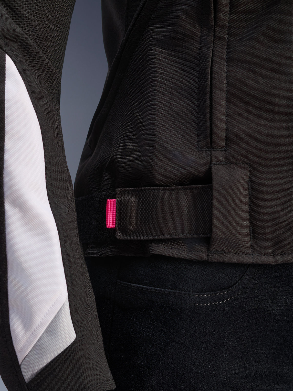 Giacca Alpinestars Stella T-GP Plus R V4, nero e bianco, primo piano del cinturino di regolazione in vita con linguetta rosa, robusta costruzione in tessuto con pannelli bianchi sulle maniche, giacca da moto da donna con vestibilità sportiva