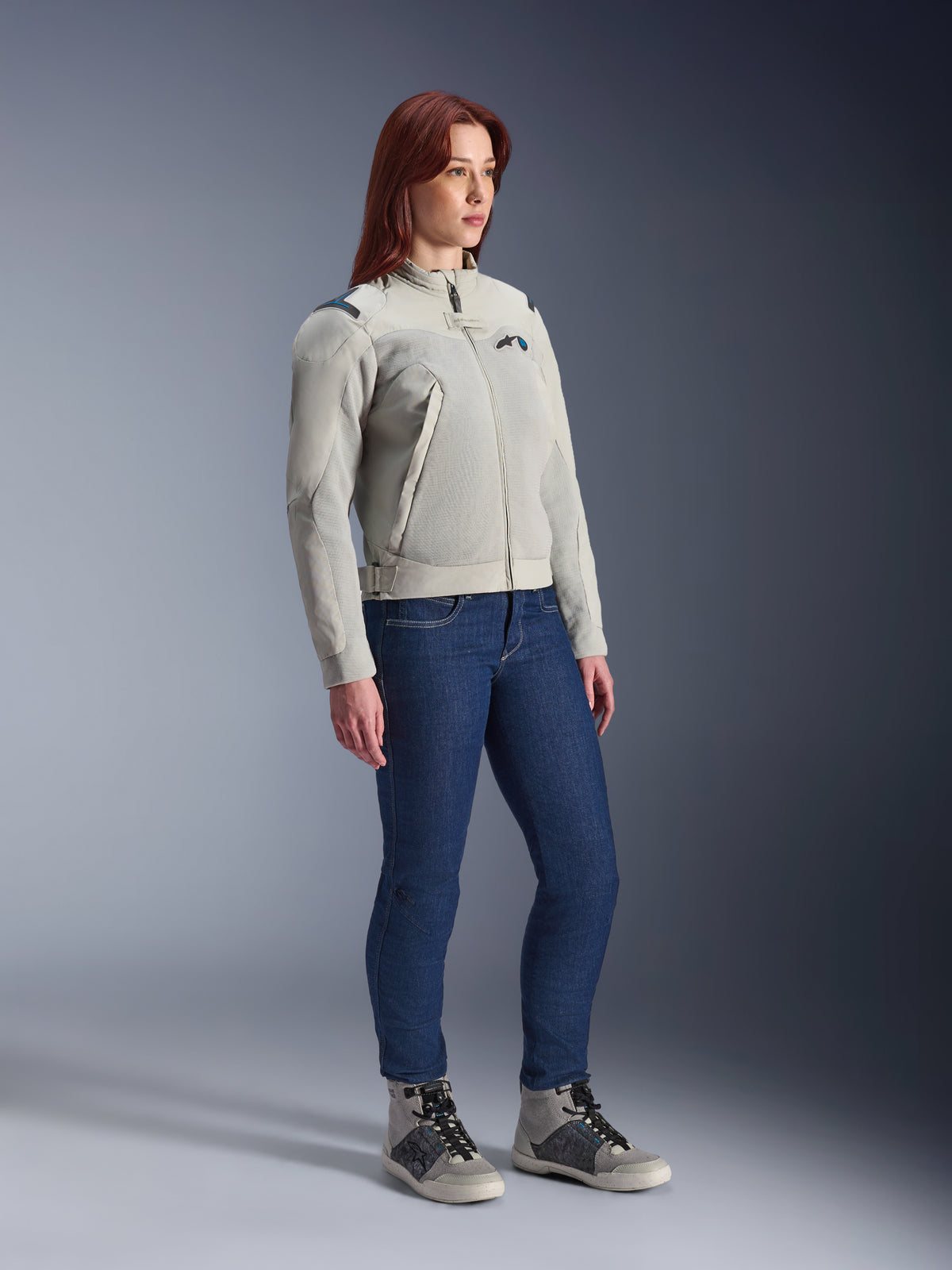 Donna che indossa la giacca Alpinestars Stella Circal-1 Air in Metal Gray e Methyl Blue, abbinata a jeans blu e scarpe da guida casual, che mette in risalto i pannelli in mesh e il design urbano minimalista della giacca.