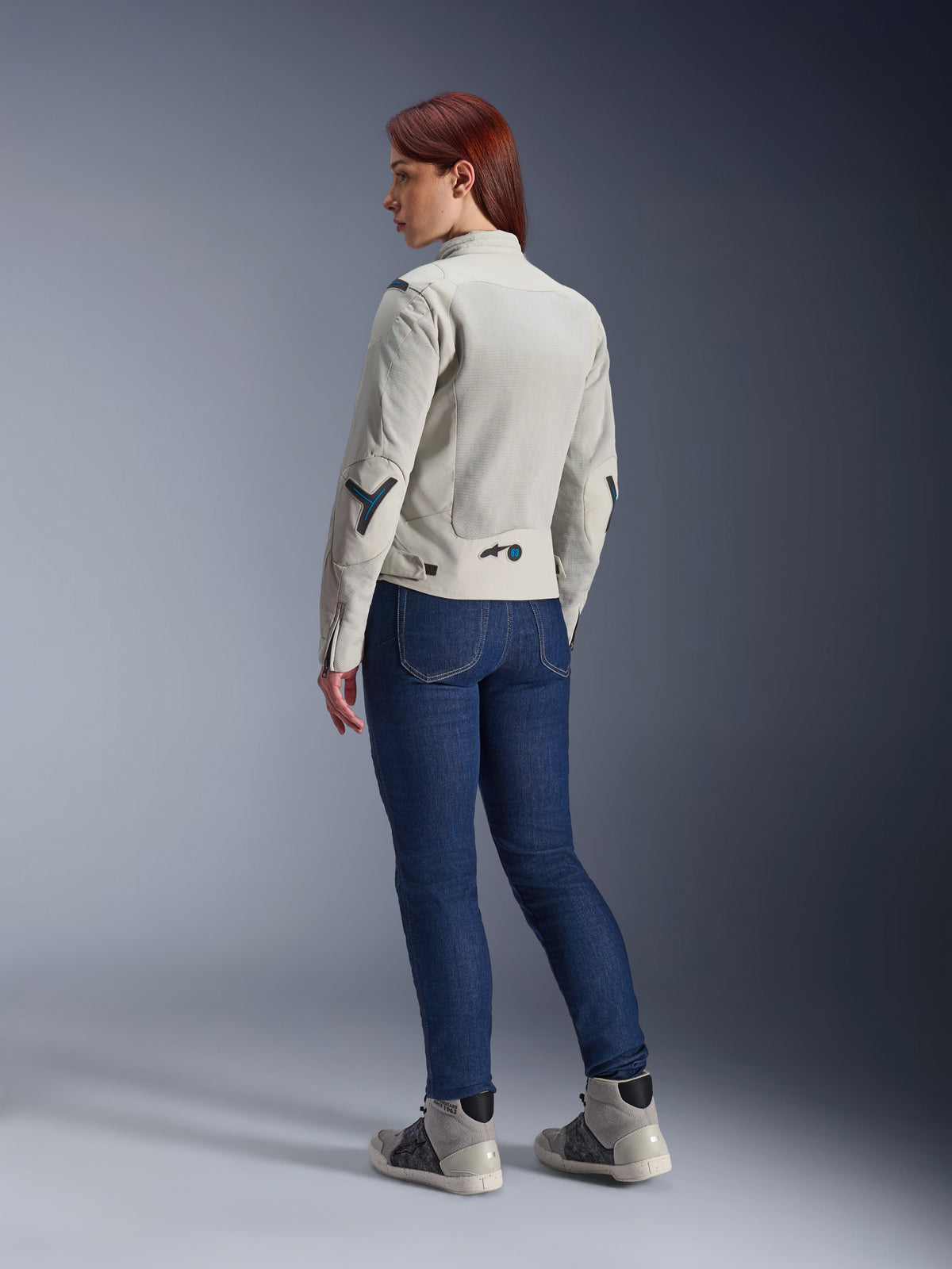 Donna che indossa la giacca Alpinestars Stella Circal-1 Air in Metal Gray e Methyl Blue, vista posteriore che mostra i pannelli in mesh per il flusso d'aria e gli accenti blu neon, abbinata a jeans da moto blu scuro e sneakers grigie, scatto lifestyle motociclistico urbano.