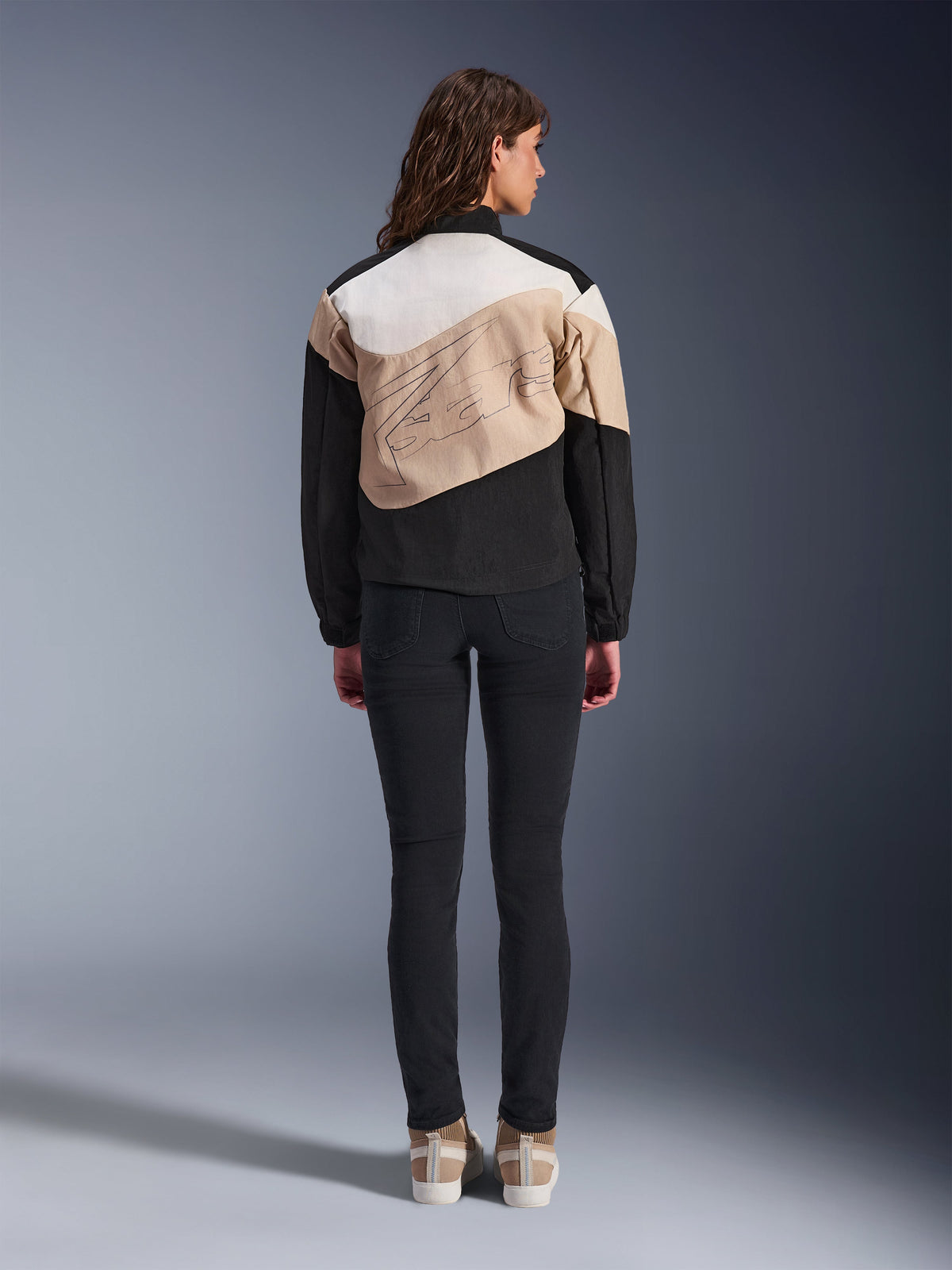 Giacca Alpinestars Stella Moshell, giacca da moto urbana da donna, vista posteriore, design asimmetrico Black Sand e Off White con profilo grafico grande, indossata con pantaloni neri slim-fit e sneaker color sabbia, abbigliamento per guida urbana