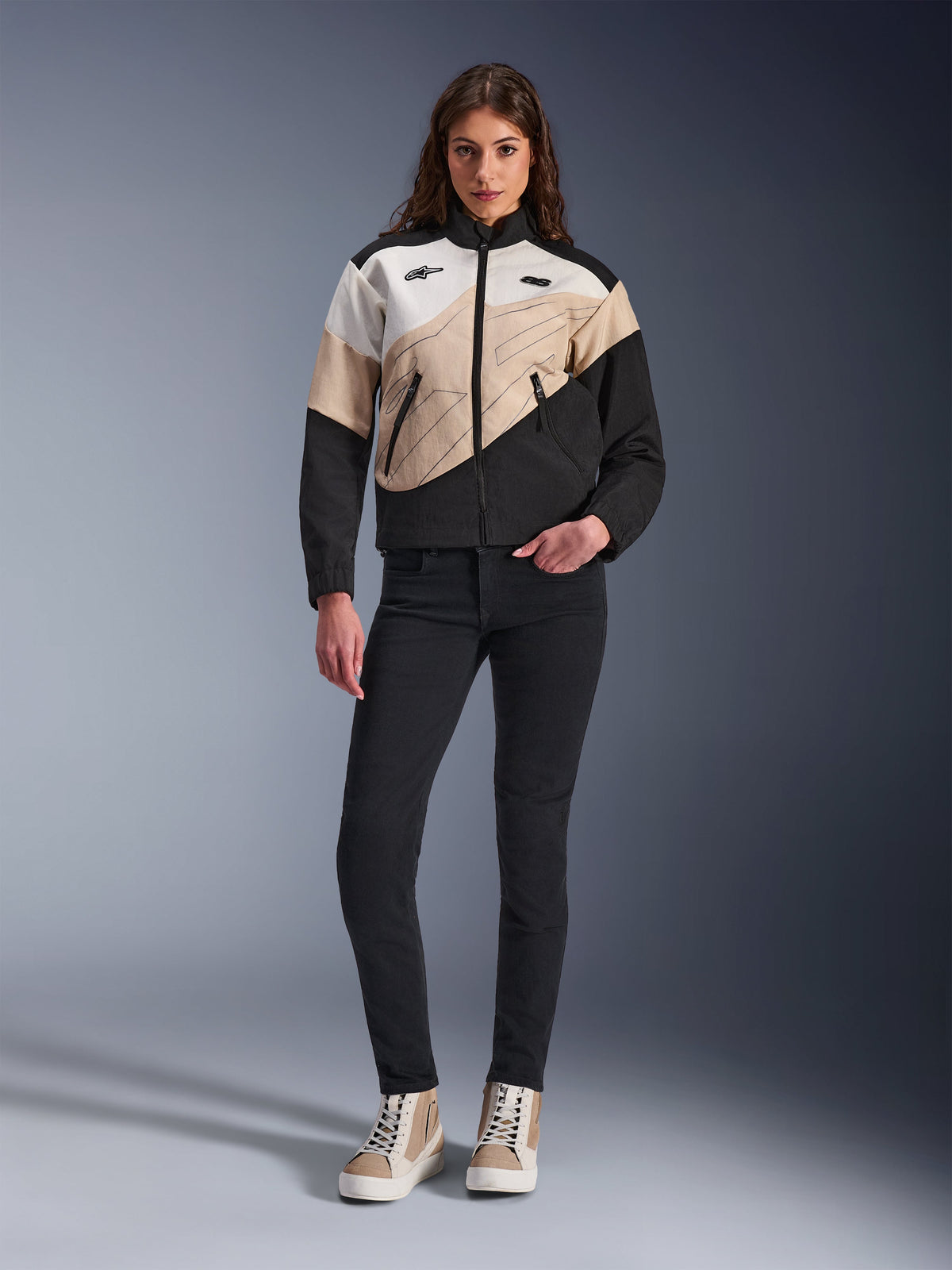 Giacca Alpinestars Stella Moshell da donna, giacca da moto urbana con design asimmetrico color-block in nero, sabbia e bianco sporco, indossata con pantaloni slim-fit neri e sneakers alte color sabbia, estetica streetwear moderna per motocicliste.