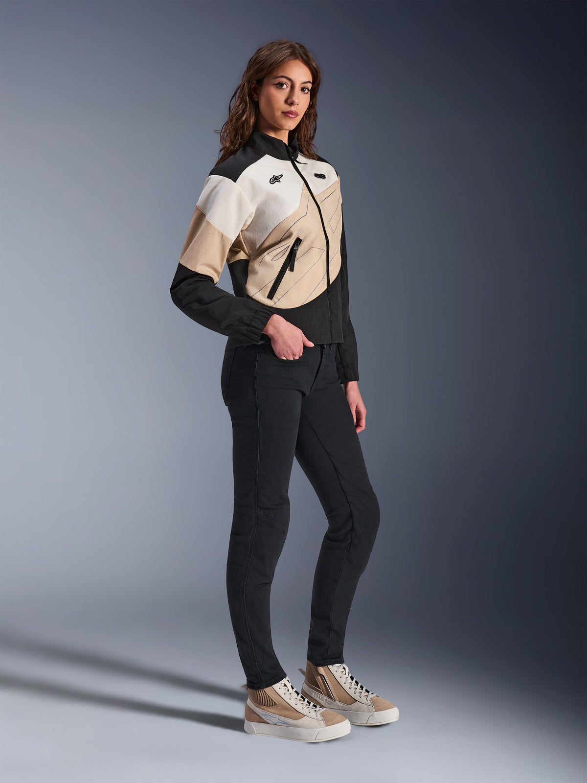 Giacca Alpinestars Stella Moshell, giacca da moto urbana da donna, nei colori Black Sand e Off White, caratterizzata da un design geometrico asimmetrico, indossata con pantaloni neri slim e sneakers color sabbia, scatto in studio a figura intera per abbigliamento da moto urbano contemporaneo.