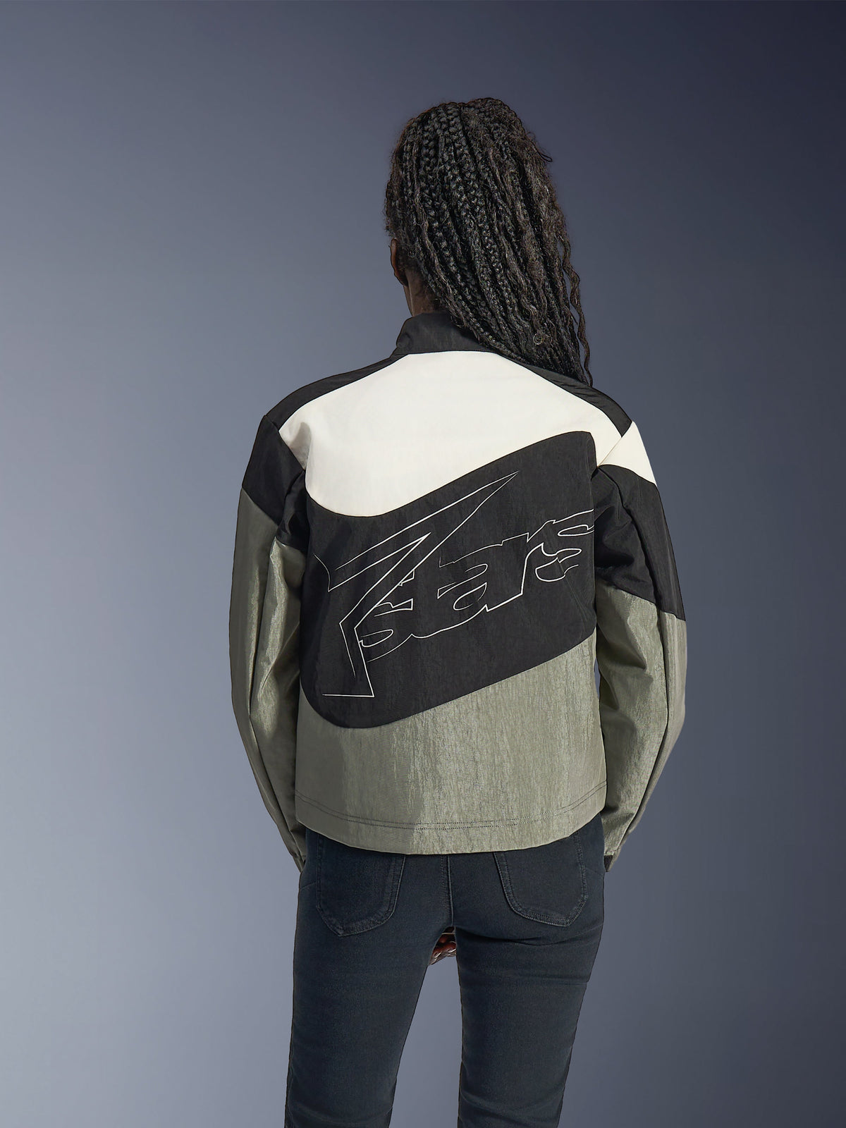 Donna vista da dietro che indossa la giacca Alpinestars Stella Moshell, giacca da moto urbana in nero, grigio e bianco sporco, caratterizzata da un grande logo profilato in bianco sul retro, abbinata a pantaloni neri, abbigliamento tecnico contemporaneo ispirato allo streetwear