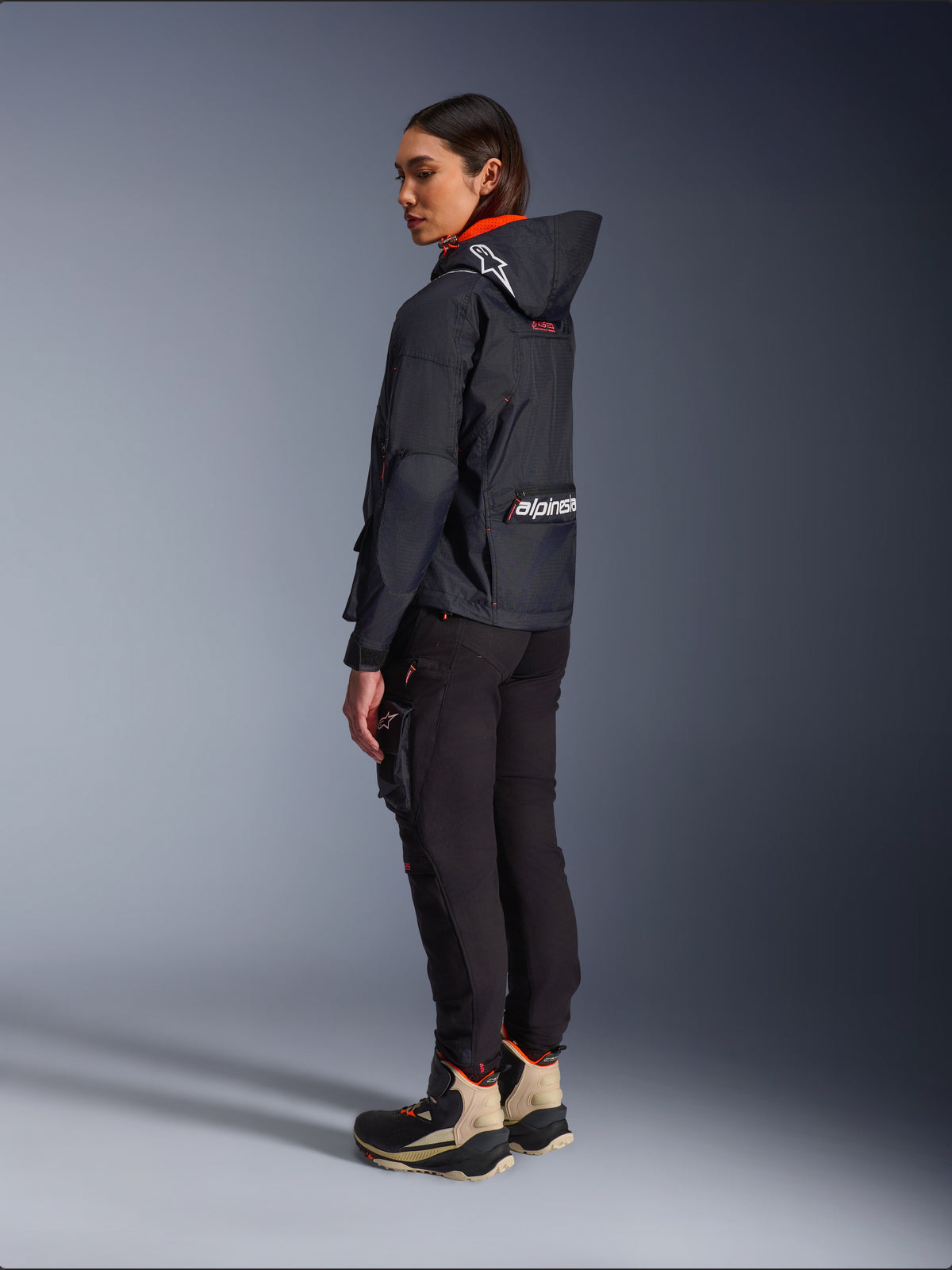 Pantaloni Alpinestars Actea da Donna, nero, moderni pantaloni da moto con vestibilità affusolata e tasca cargo sulla coscia, mostrati da una prospettiva laterale-posteriore su una modella che indossa una giacca scura con cappuccio e stivali da moto, stile urban riding.